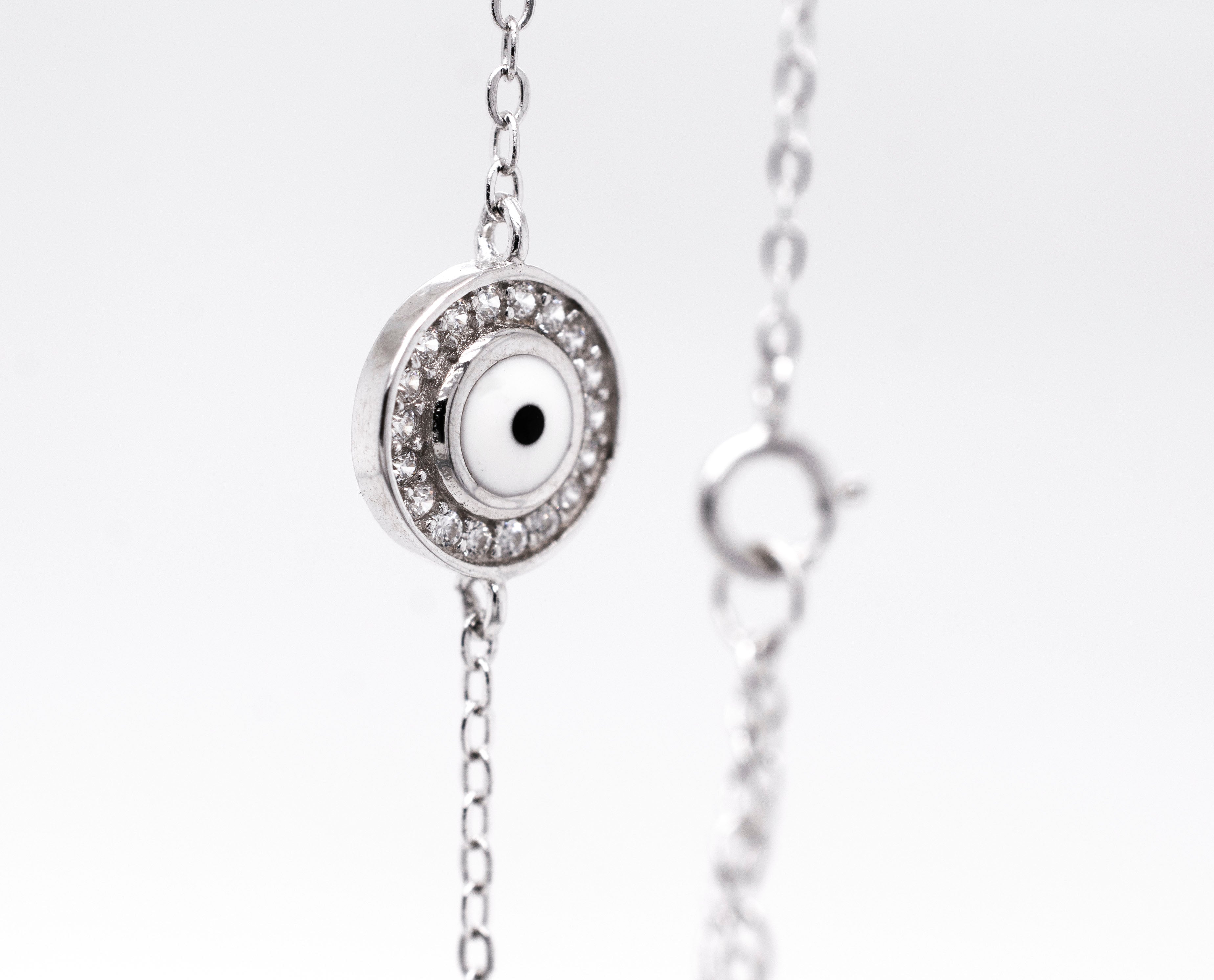 Close-up of a sterling silver Delicate Cubic Zirconia Evil Eye Bracelet with a round, eye-like charm surrounded by small, clear cubic zirconia gemstones. The clasp of the bracelet is visible in the background.
