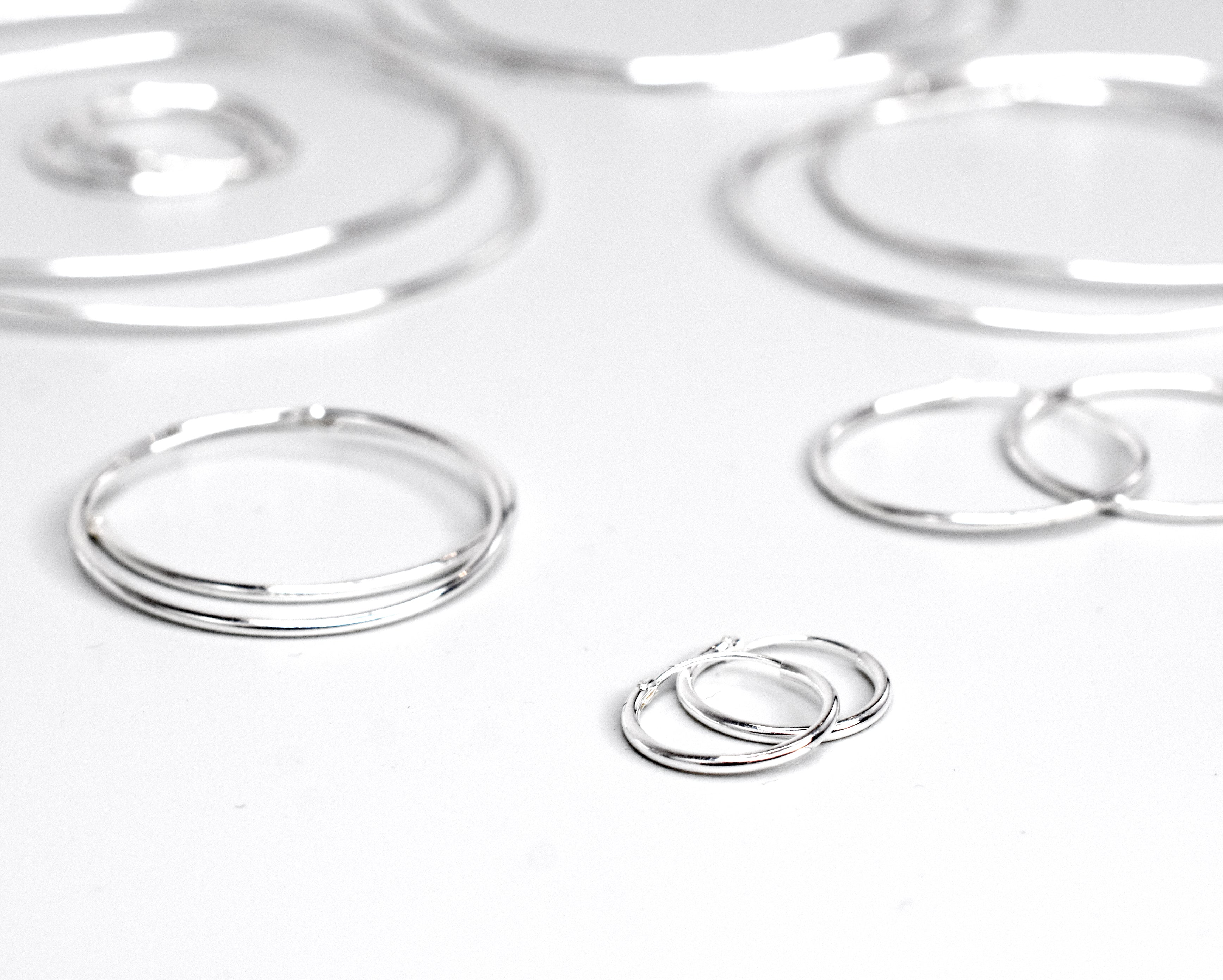 Displayed on a white surface, the 1.2mm Infinity Hoops feature an elegant infinity hoop design in varying sizes from small to large. Each pair of these silver hoop earrings is meticulously crafted from .925 Sterling Silver, adding a touch of sophistication to any ensemble.