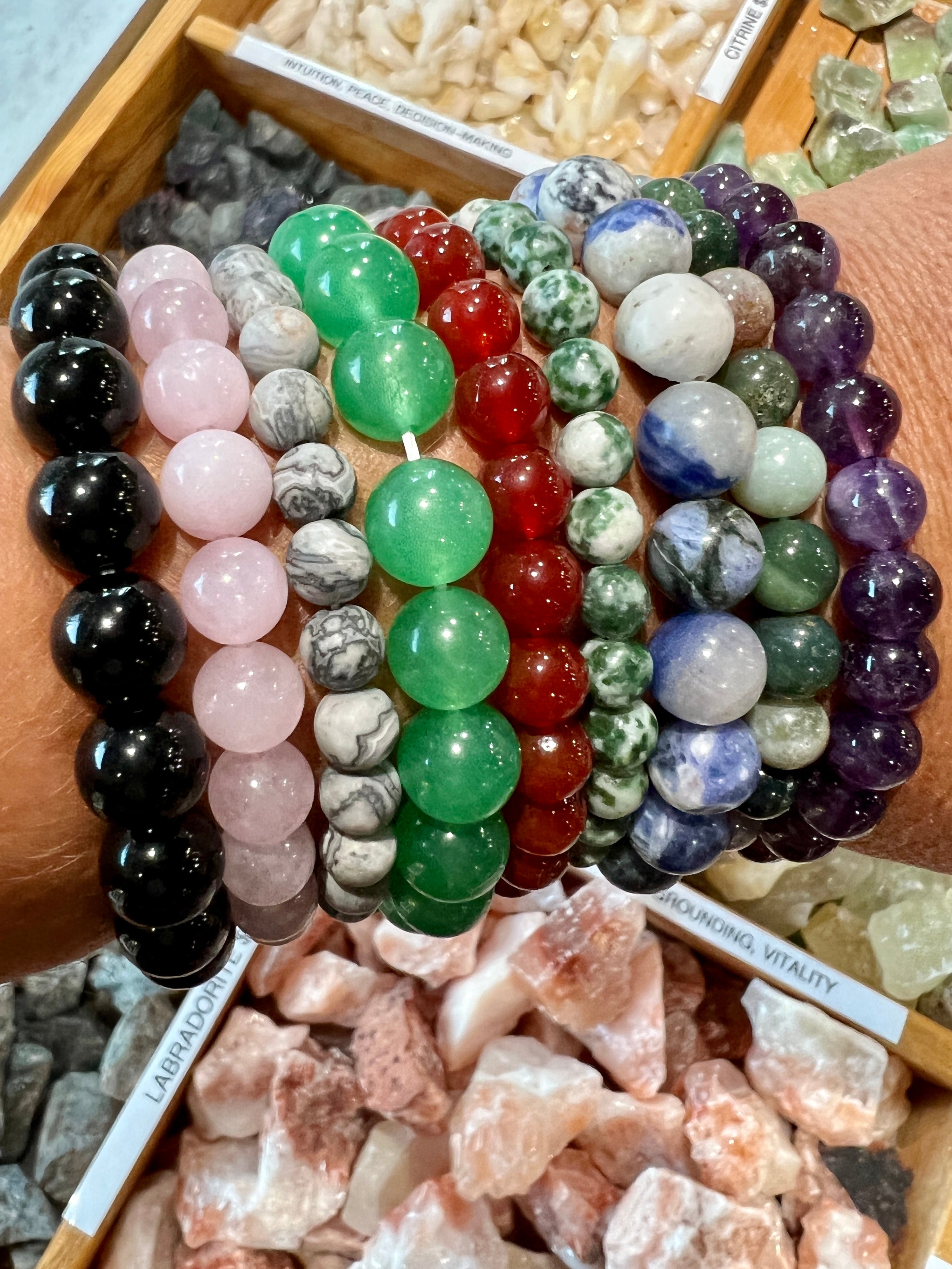 Various 4mm Beaded Stone Bracelets displayed on a wrist, above trays of assorted crystal stones, with informational labels visible.