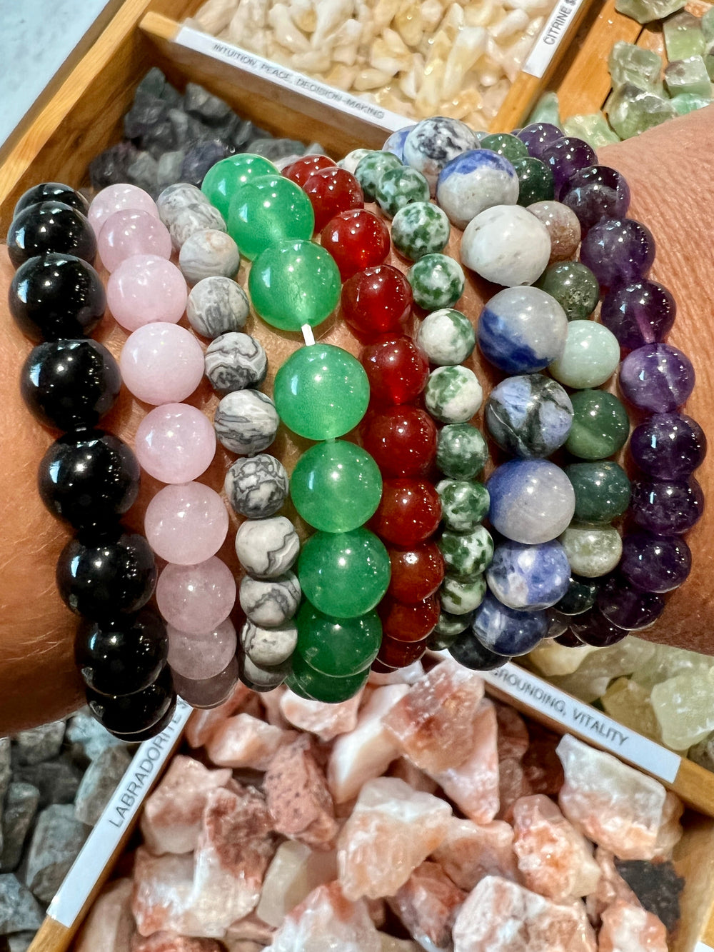Various 4mm Beaded Stone Bracelets displayed on a wrist, above trays of assorted crystal stones, with informational labels visible.