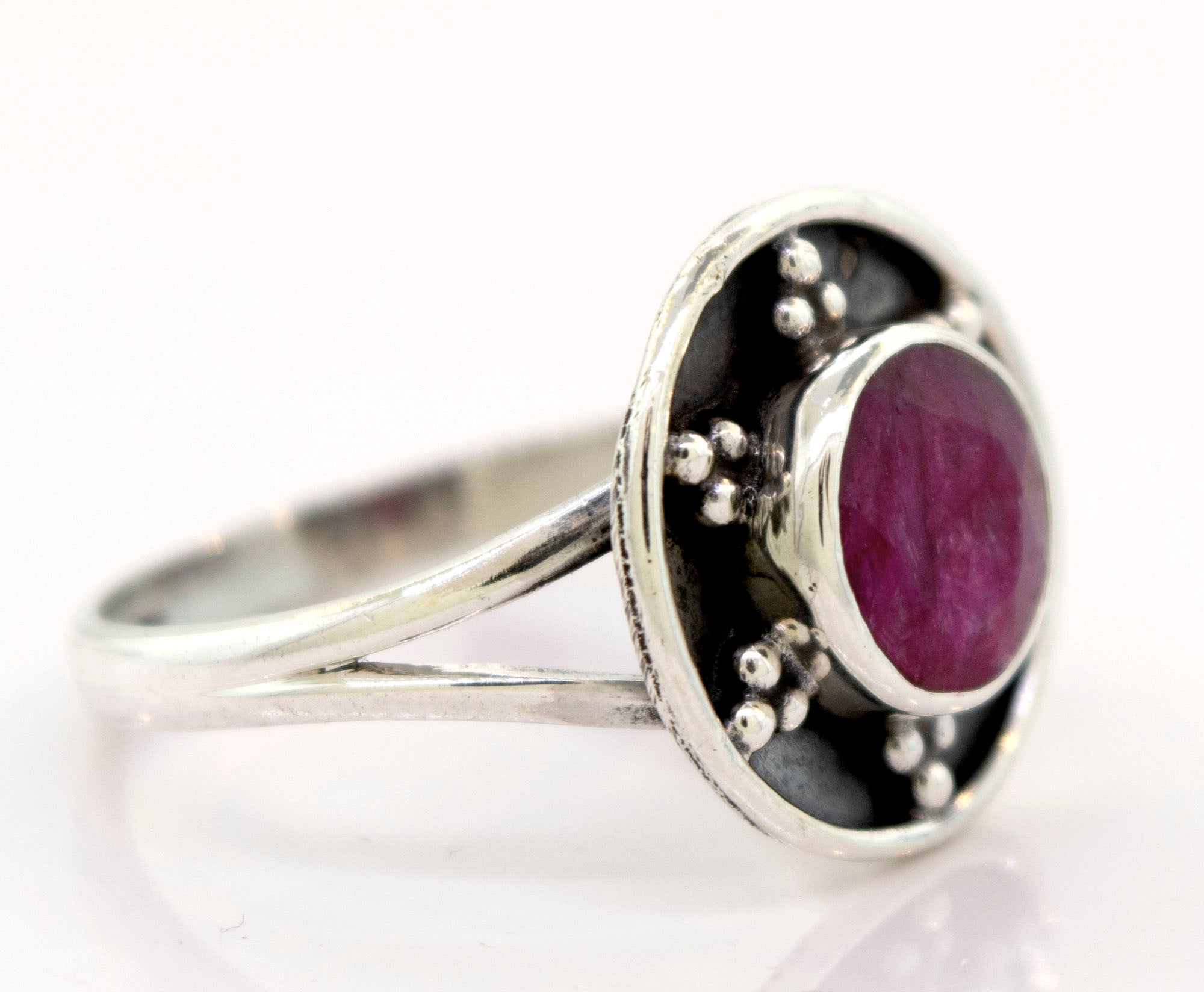 A Gemstone Ring With Unique Oxidized Design with a round, red gemstone set in the center, surrounded by small decorative metal beads. The band splits into two as it approaches the setting, creating an elegant piece of gemstone jewelry.