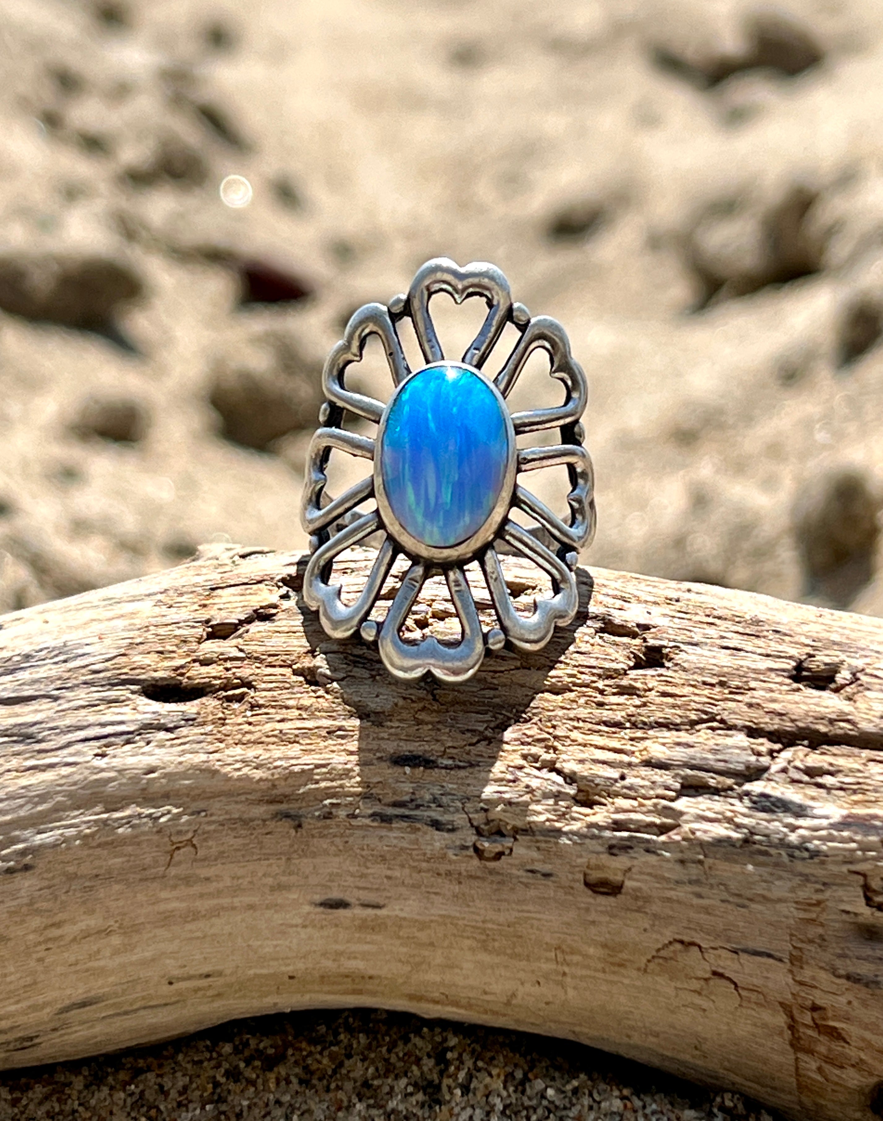 A Super Silver American Made Opal Flower Ring with Heart Shaped Petals, resting on top of a piece of driftwood.