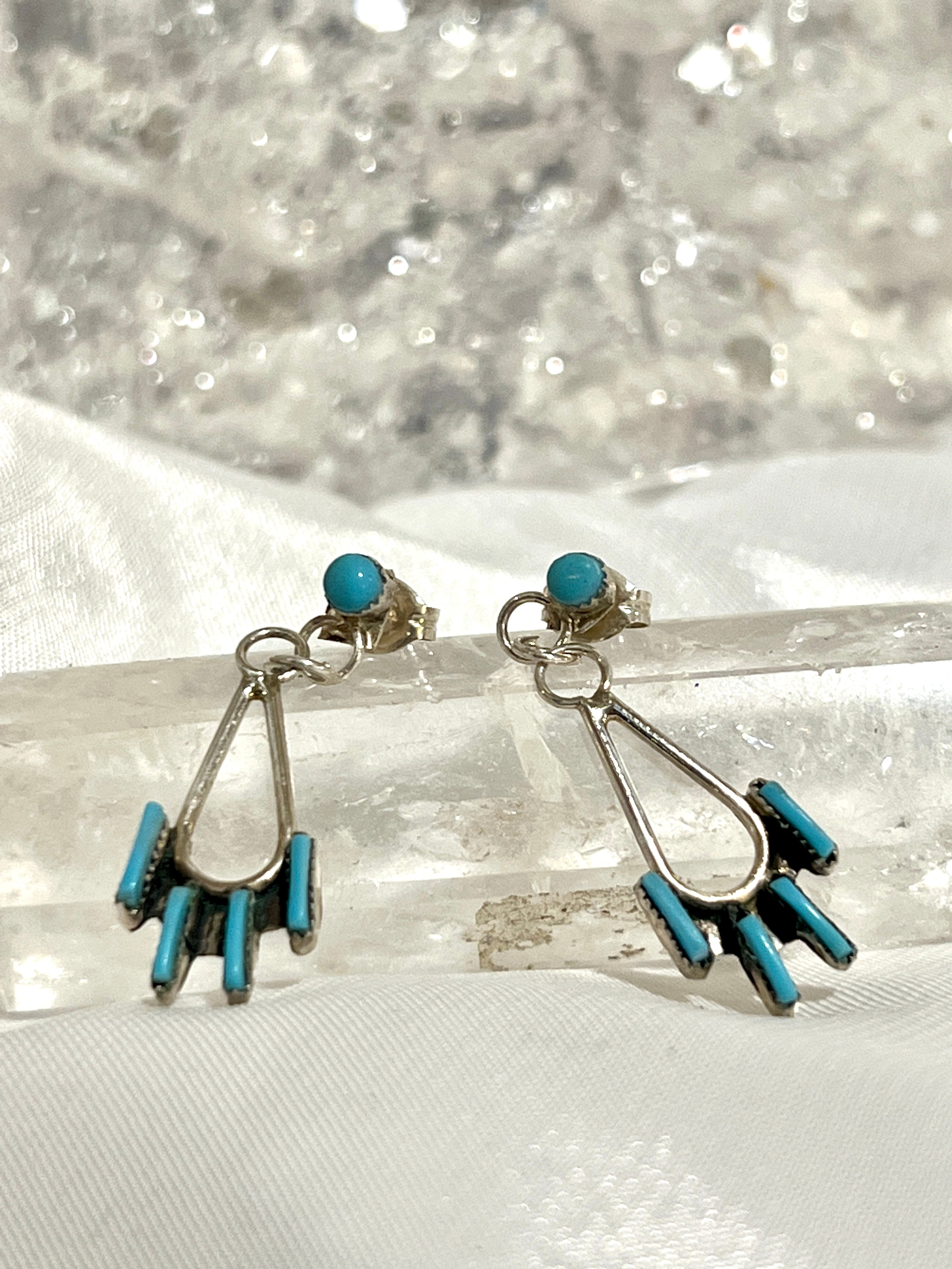 A pair of Super Silver Delicate Needlepoint Zuni Turquoise Earrings sitting on top of a crystal.