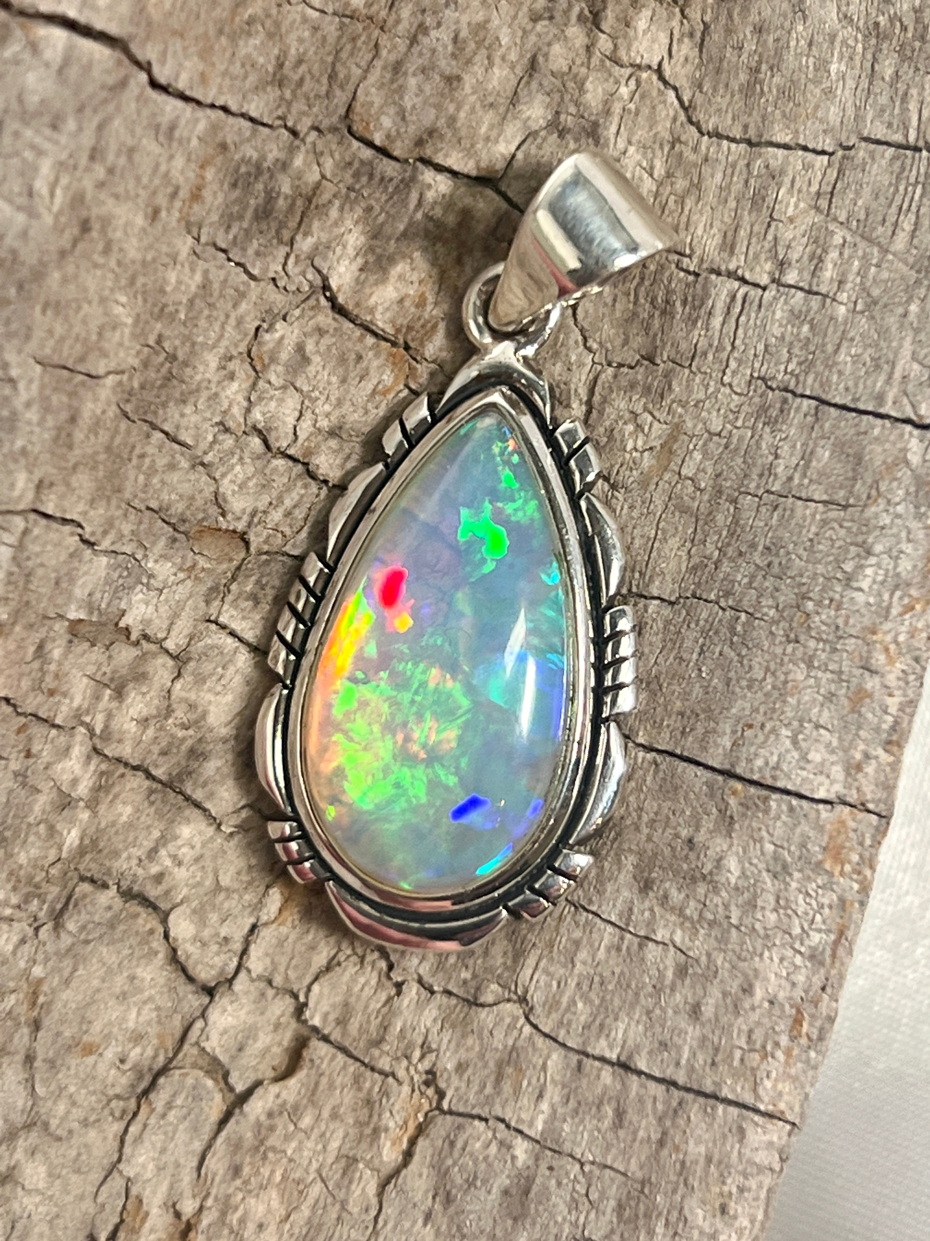 A stunning Super Silver Ethiopian Opal Teardrop Shaped Pendant with Detailed Setting, serving as a captivating statement piece, adorning a rustic piece of wood.