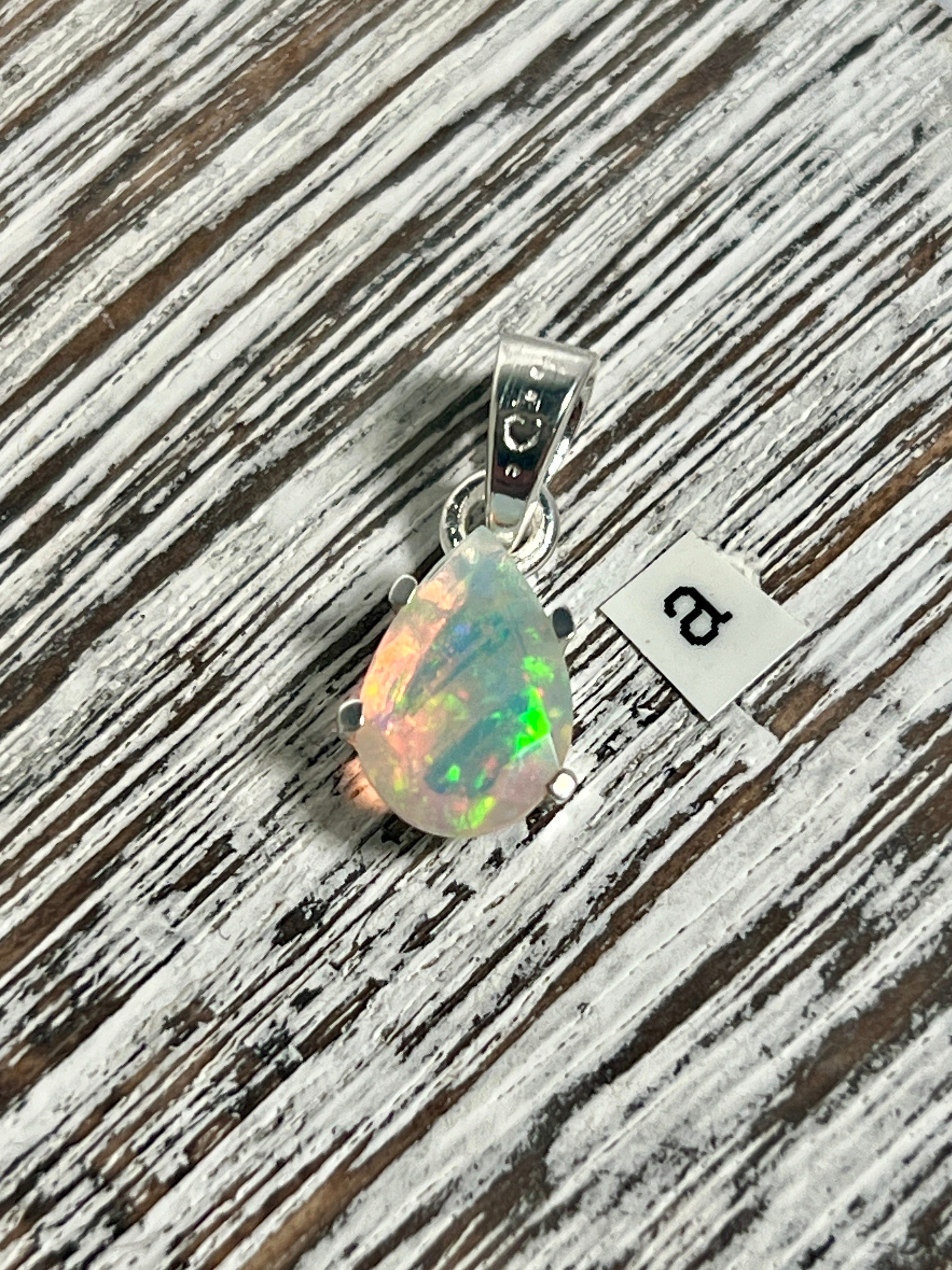 A Dainty Prong Set Facet Cut Teardrop Shaped Ethiopian Opal Pendant from Super Silver resting on a wooden table.