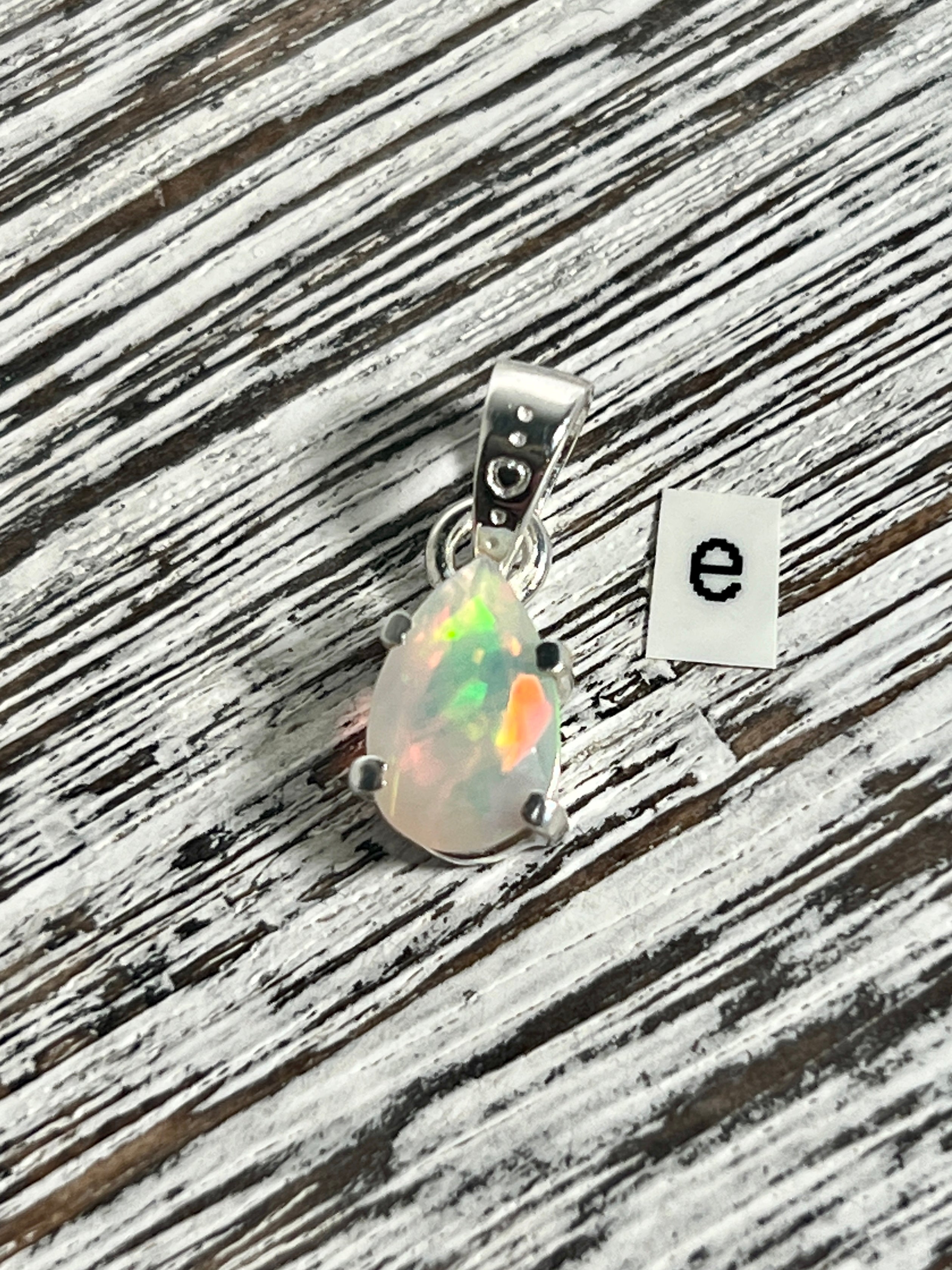 A Dainty Prong Set Facet Cut Teardrop Shaped Ethiopian Opal Pendant adorned with a stunning Ethiopian opal and featuring the letter e, produced by Super Silver.