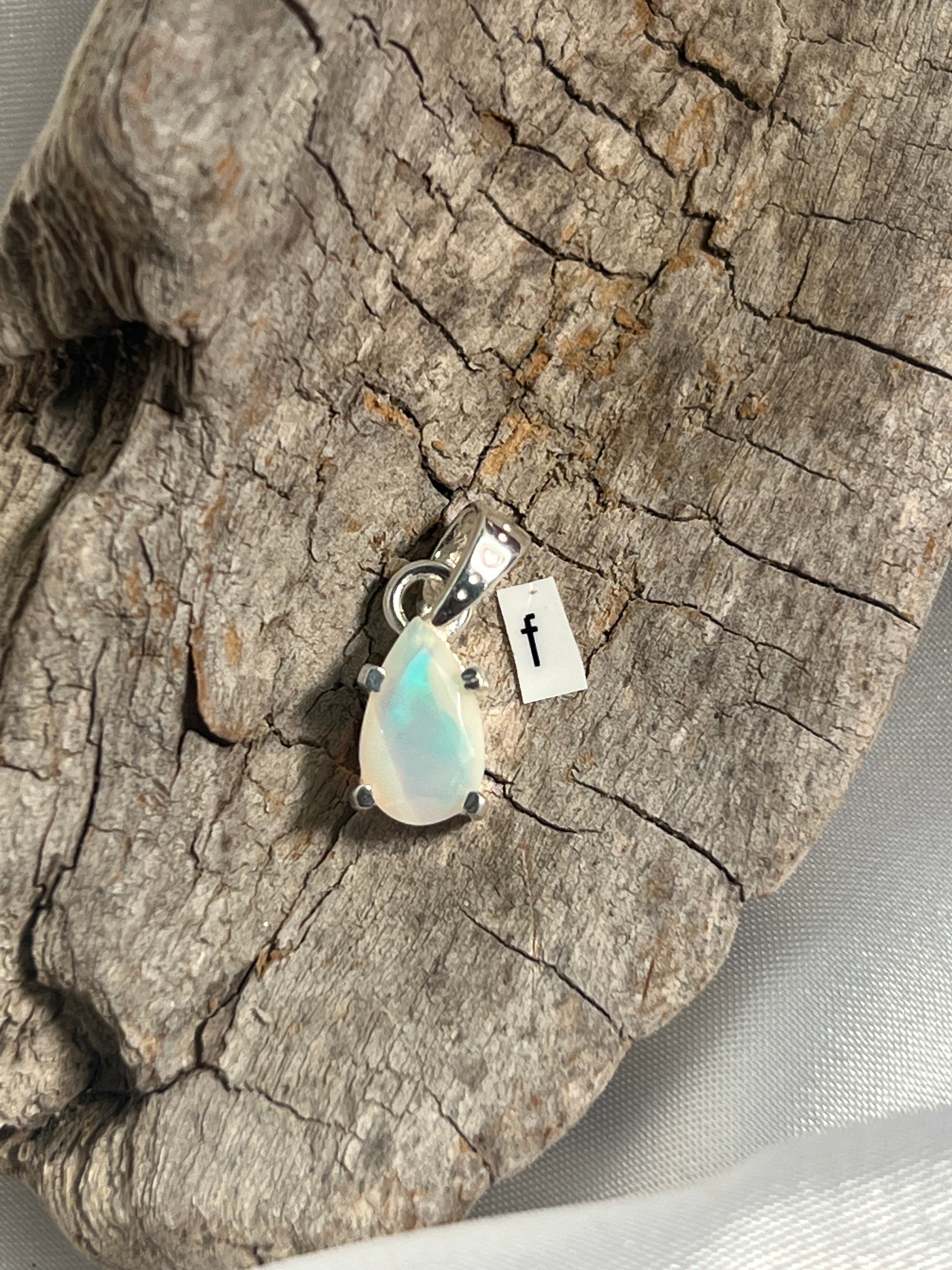 A stunning Dainty Prong Set Facet Cut Teardrop Shaped Ethiopian Opal Pendant on a piece of wood with a Super Silver accent.