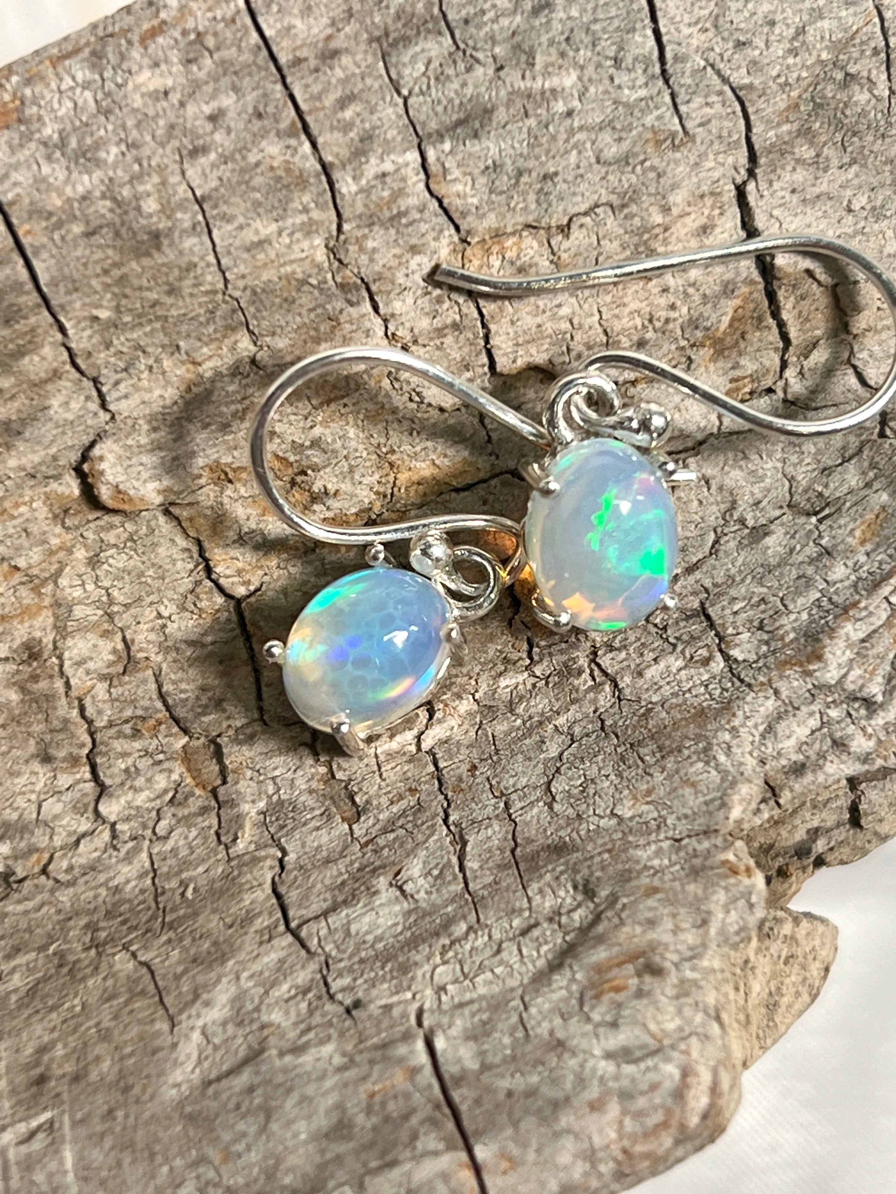 A pair of Super Silver Vibrant Oval Ethiopian Opal Earrings, with vibrant hues and natural brilliance, delicately displayed on a piece of wood.