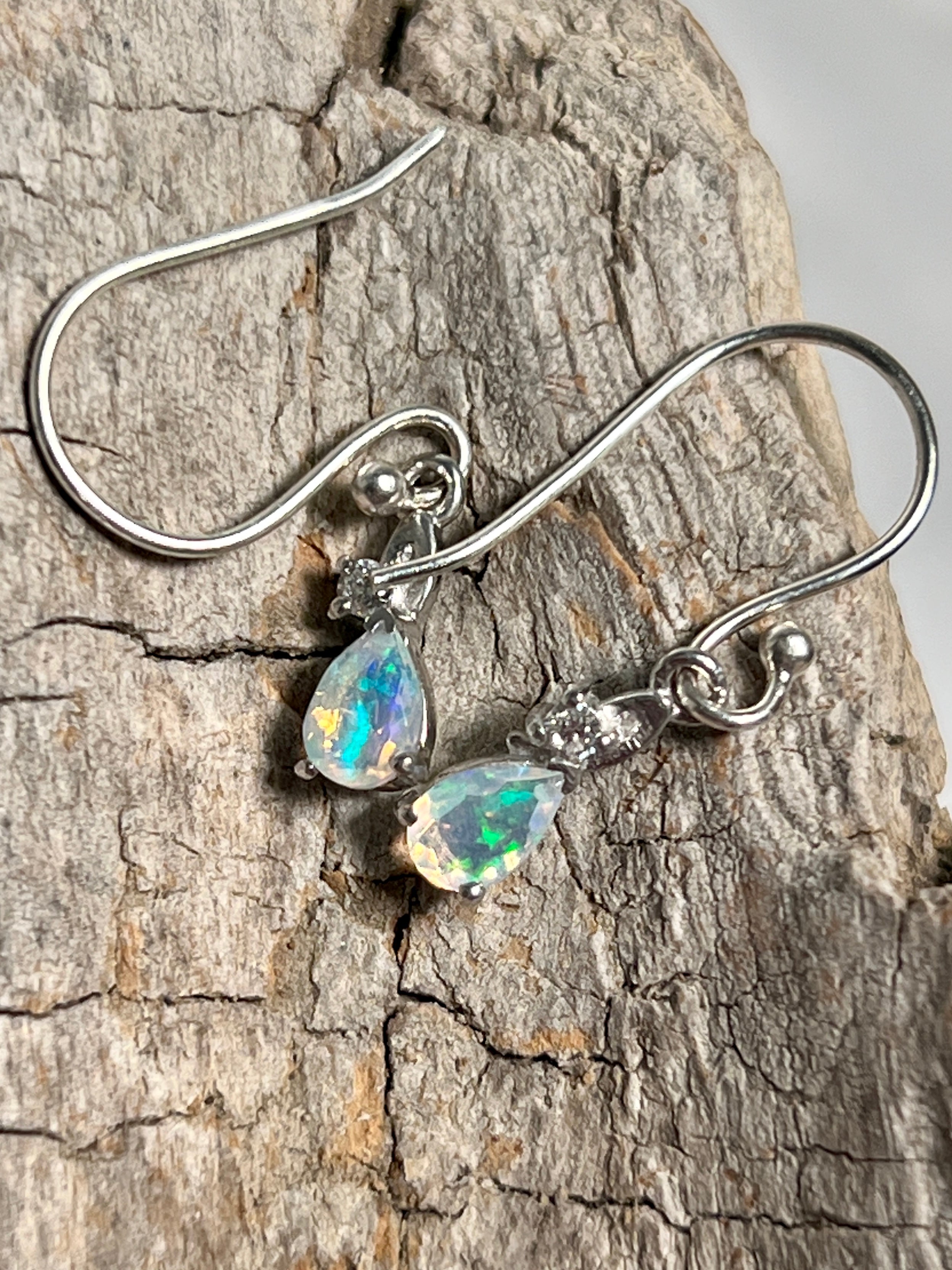 A pair of Tiny Teardrop Ethiopian Opal Earrings sparkling on a piece of wood, by Super Silver.