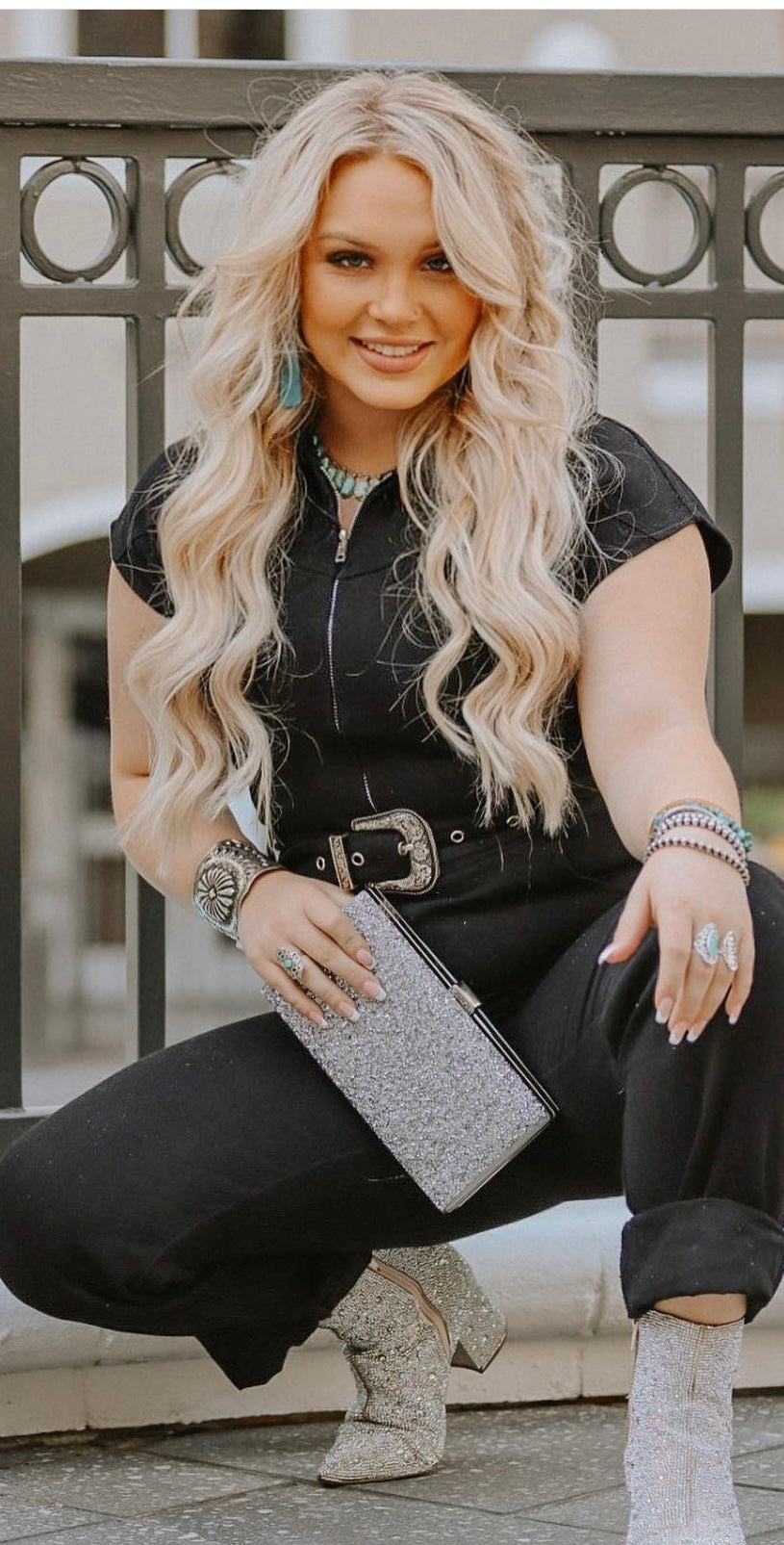 A person with long blonde hair is wearing a black outfit, sterling silver boots, and a glittery clutch. Adorned with a Handcrafted Silver Concho Cuff, they are posing in front of a railing, smiling at the camera.