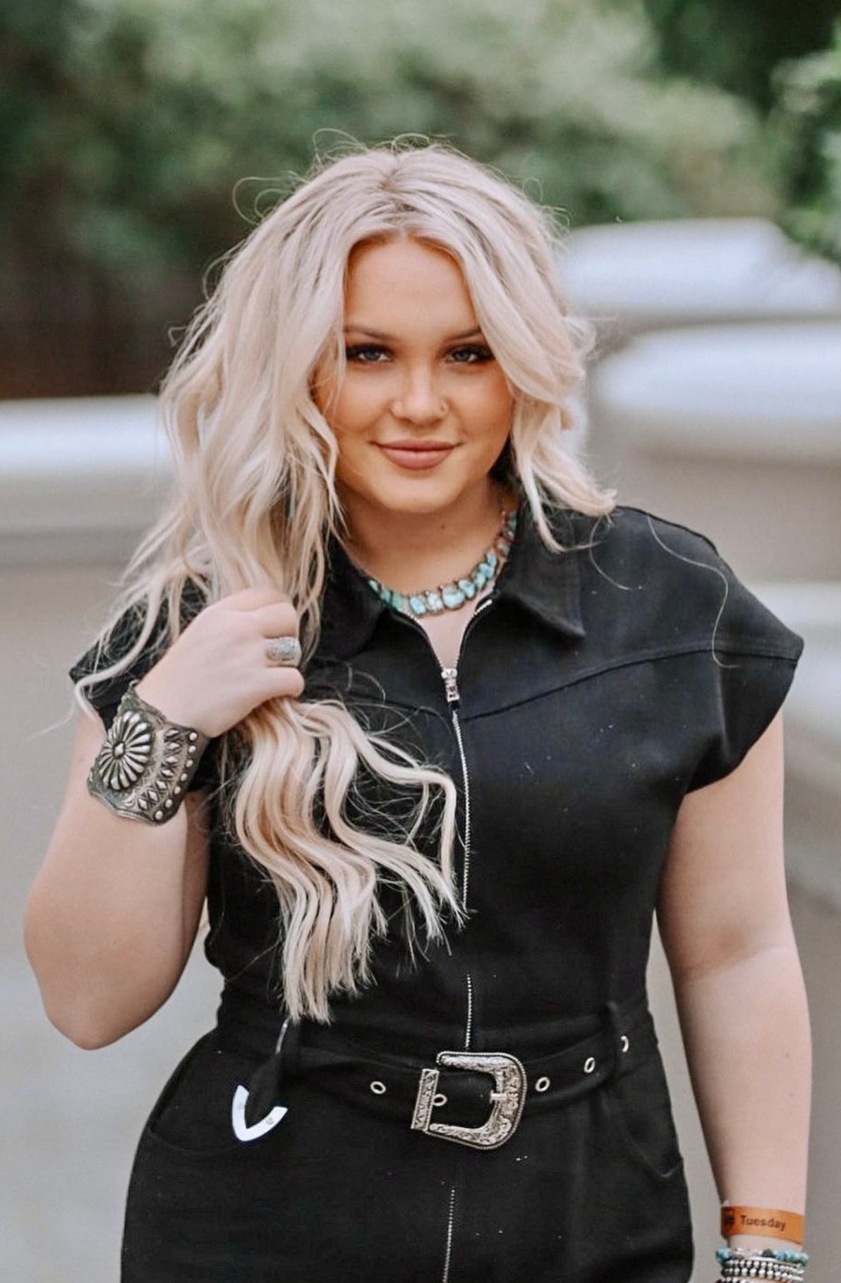 A woman with long blonde hair wearing a black outfit, accessorized with a Handcrafted Silver Concho Cuff, necklace, bracelet, and rings, stands outdoors.