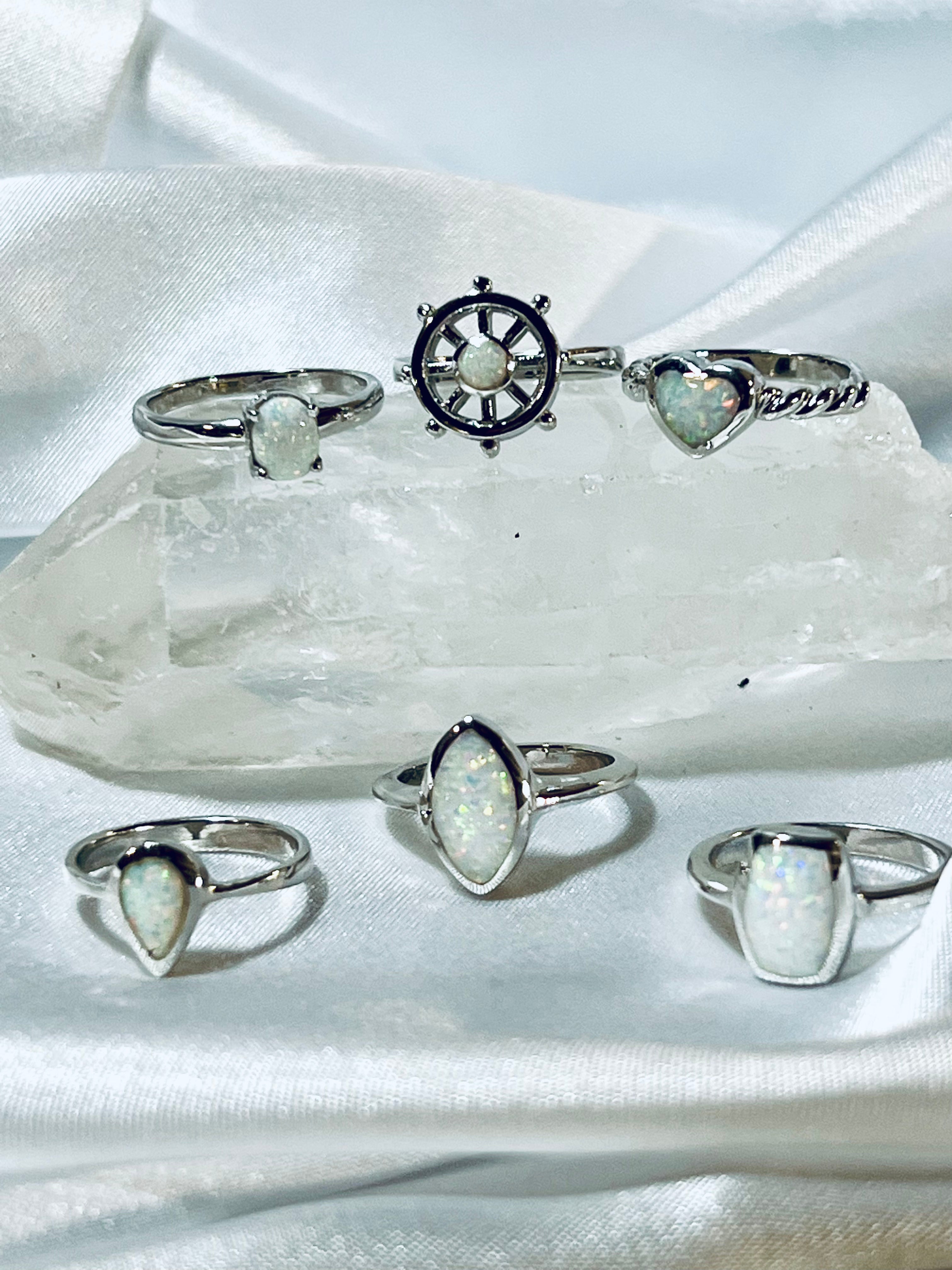 A collection of rhodium-plated silver rings with Rectangular Lab Opal Rings displayed on a crystal atop a draped white fabric.
