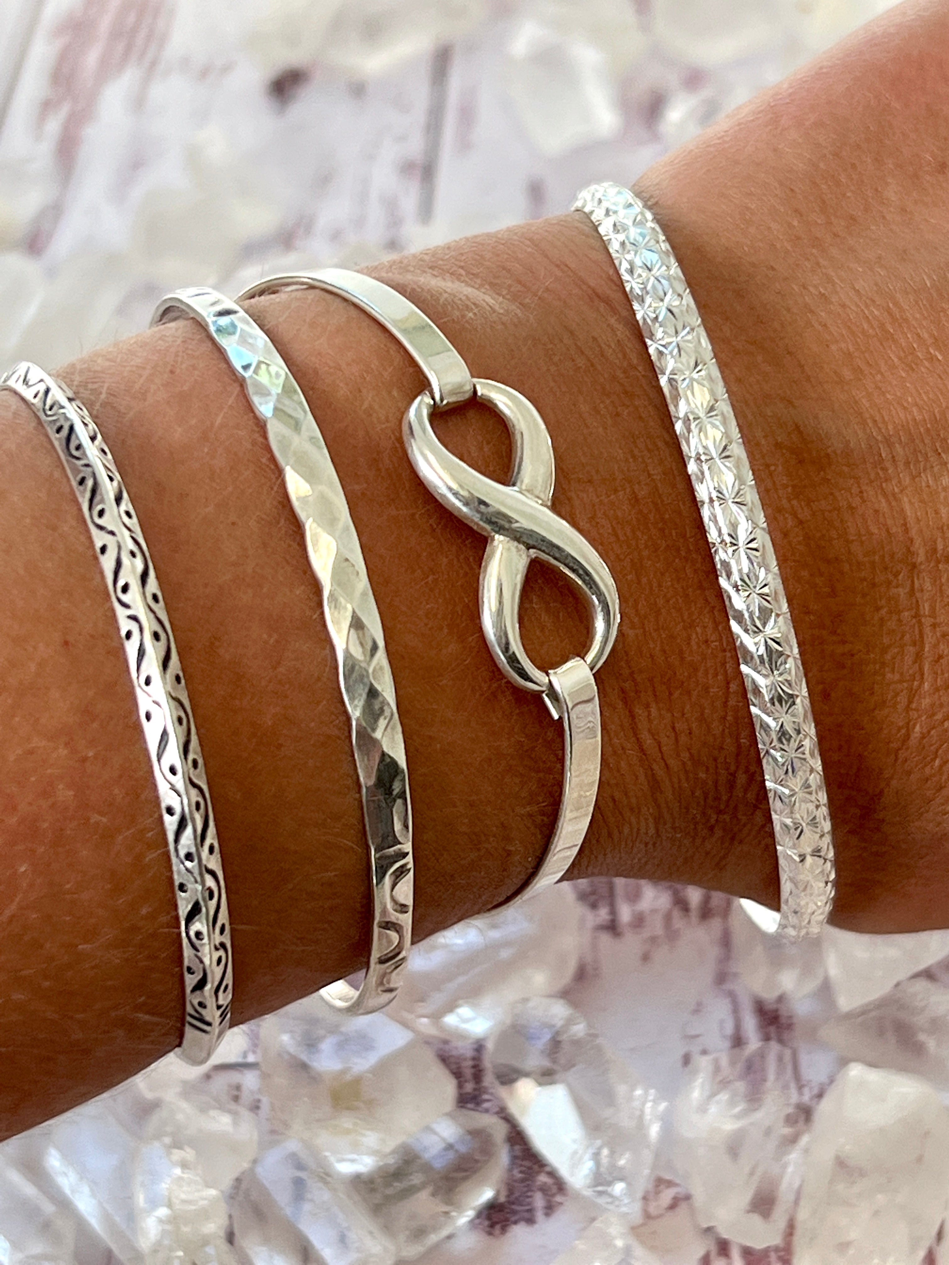 A woman's wrist adorned with three sleek Super Silver bracelets, featuring an elegant Infinity Bracelet and secured with a latch clasp.