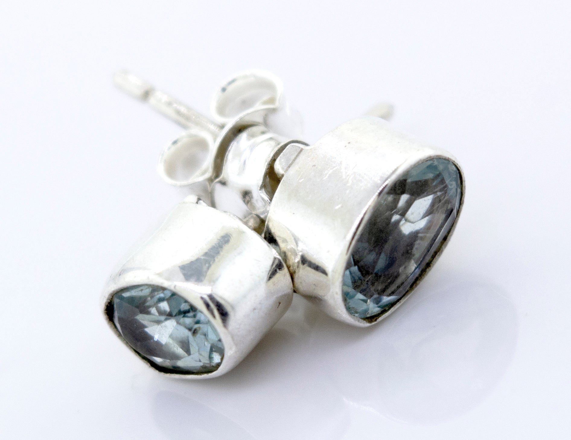 A pair of Beautiful Oval Faceted Cut Blue Topaz Studs by Super Silver with a silver setting on a white surface.