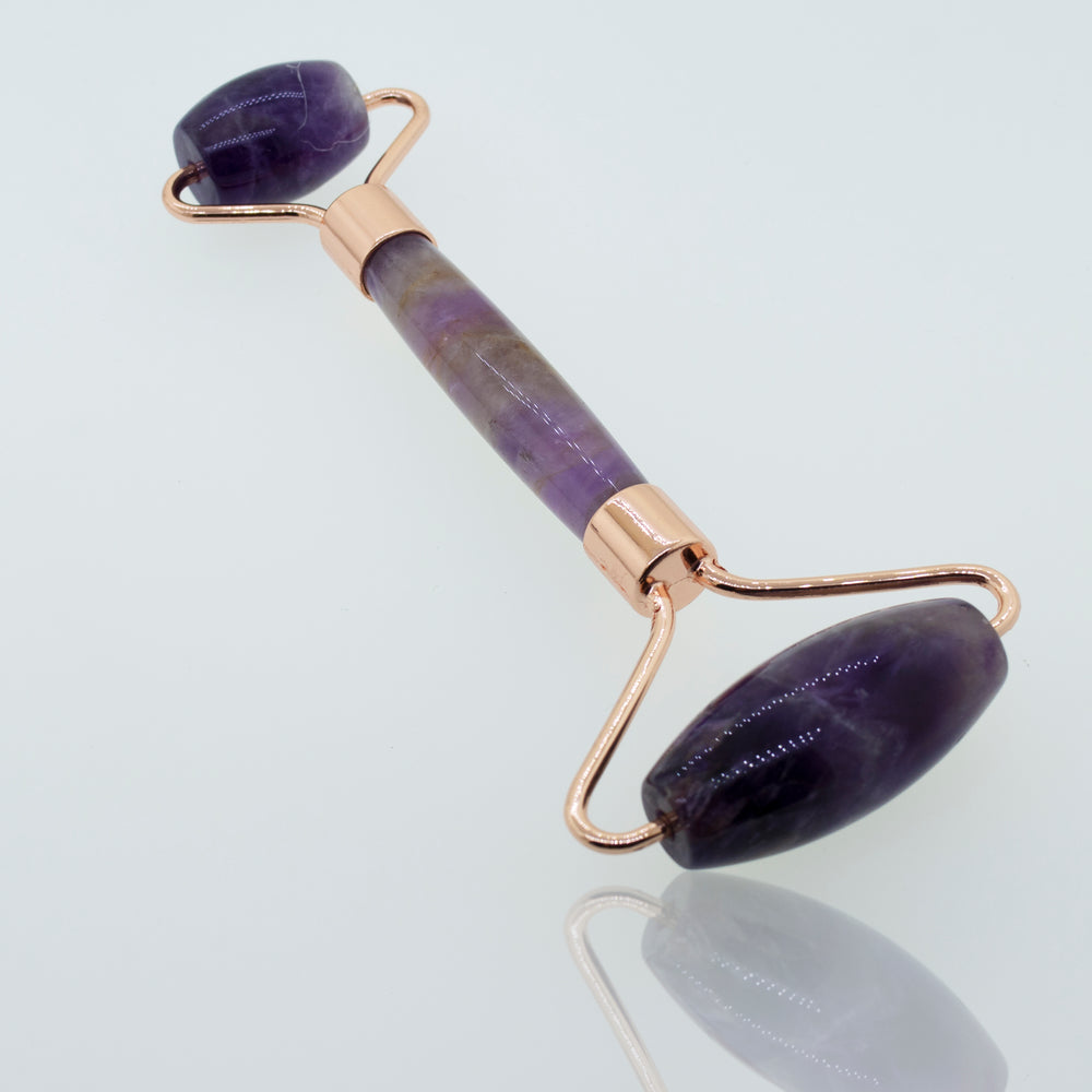
                  
                    A Stone Face-Roller with a rose gold handle and two polished purple gemstones, positioned on a reflective surface.
                  
                