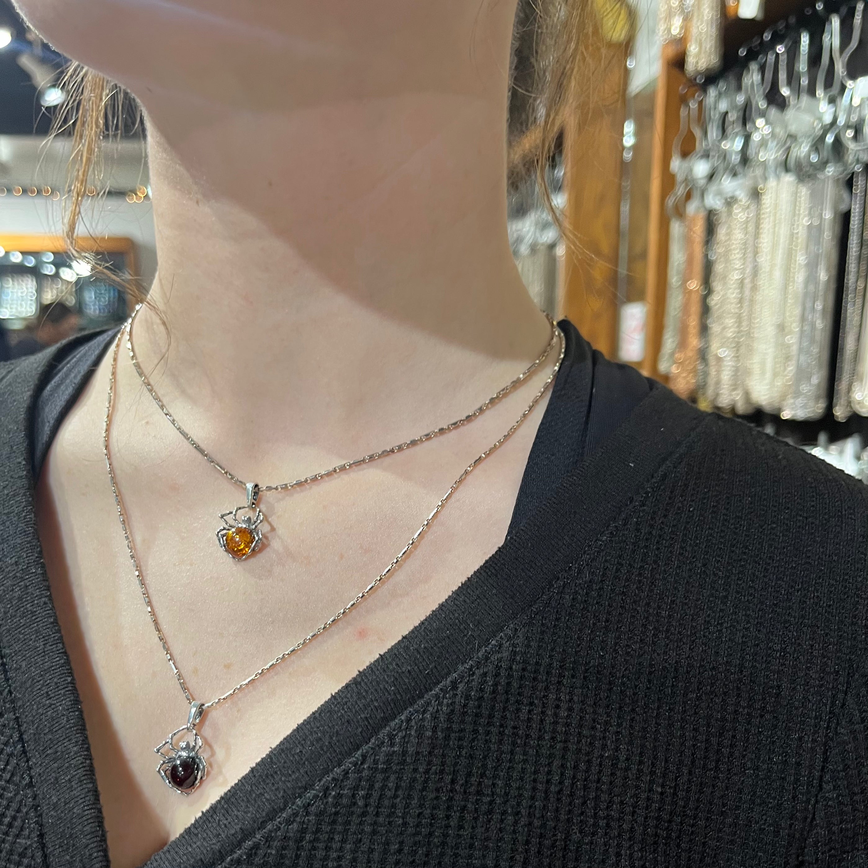 A woman is standing in a jewelry store with two necklaces, showcasing a mesmerizing Delicate Baltic Amber Spider Pendant from Super Silver and an exquisite amber stone. The scene emanates a bewitching aura, evoking a captivating witch.