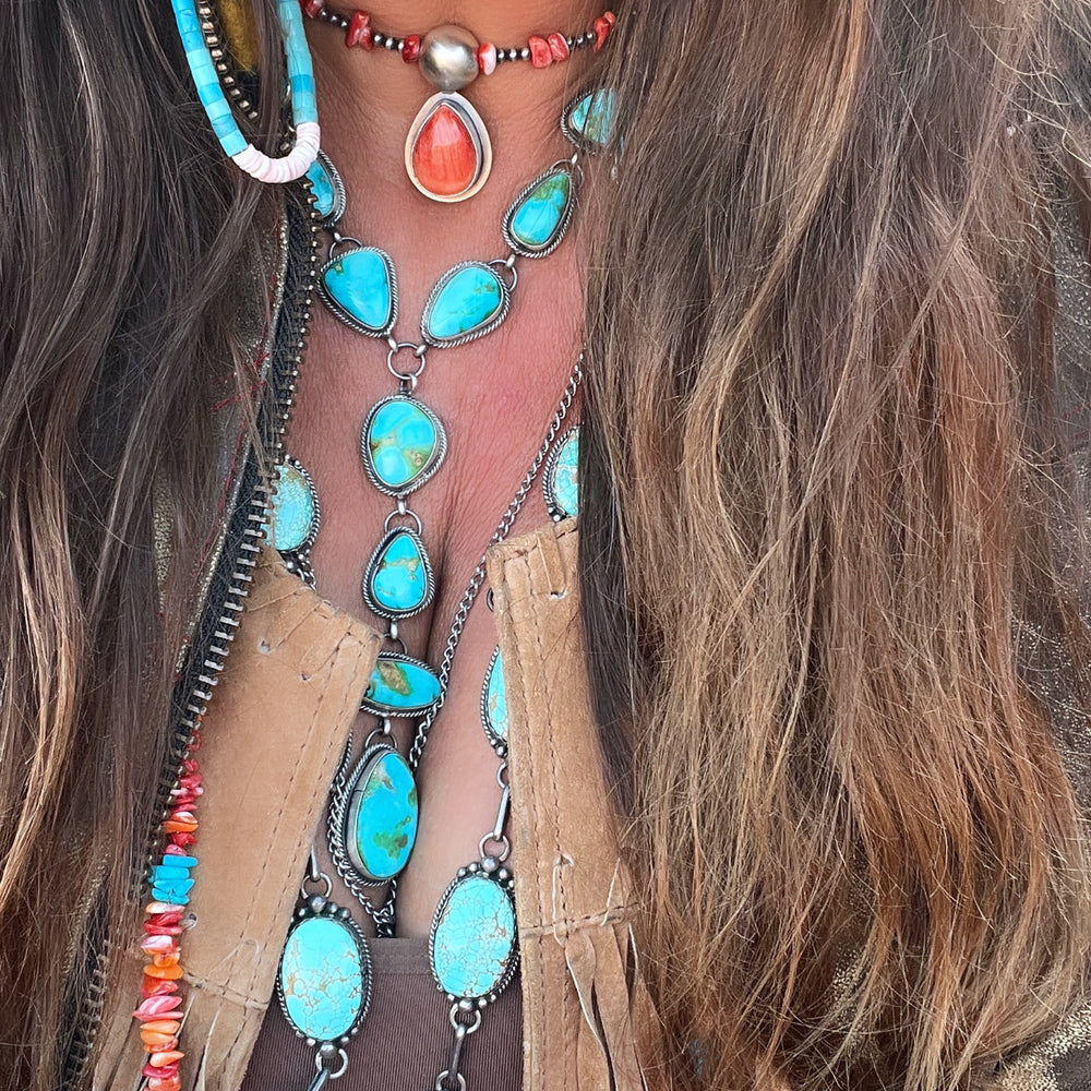 
                  
                    A person wearing multiple turquoise and coral stone jewelry pieces, a fringed top, a feather earring, and a Wrap Around Spiny Oyster Shell Choker Necklace crafted by Native American artisans.
                  
                