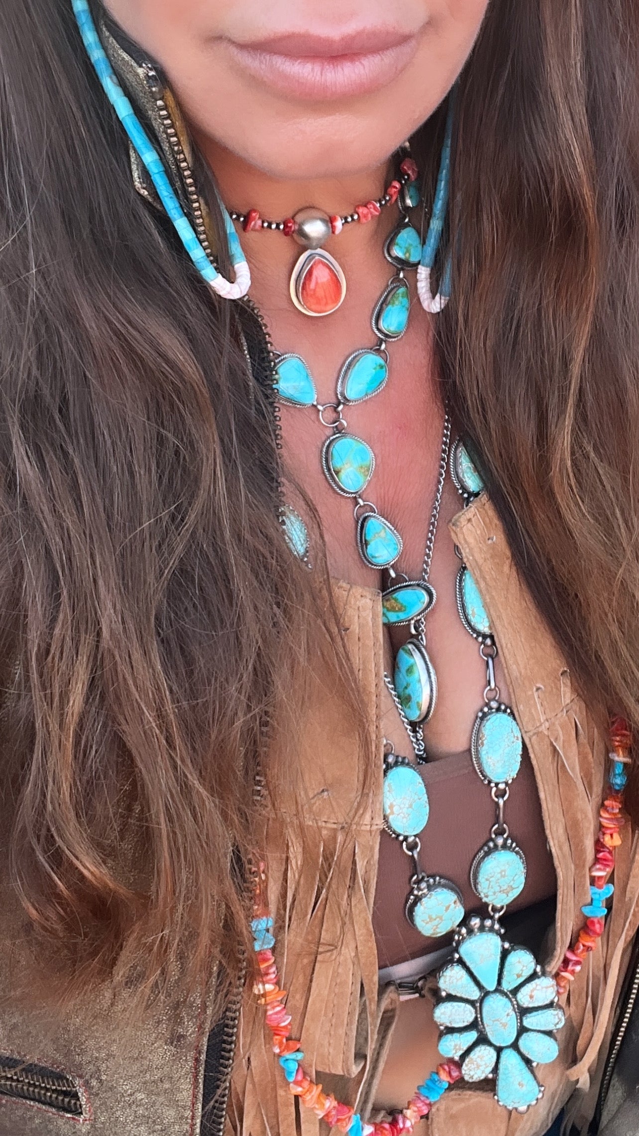 A person wearing a fringed tan leather jacket, a brown top, and layered turquoise jewelry crafted by Native American artisans, including necklaces, earrings, and a Wrap Around Spiny Oyster Shell Choker Necklace.