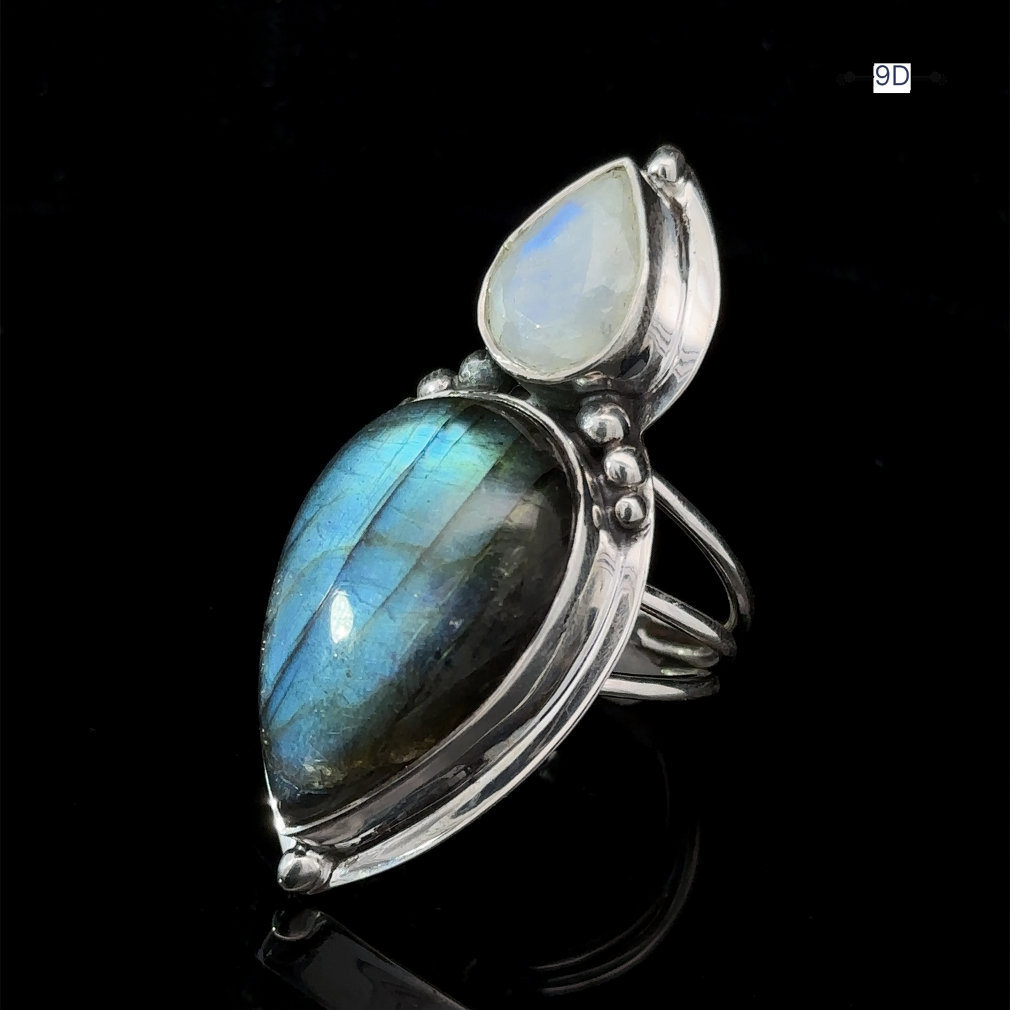 
                  
                    The Heavy Labradorite And Moonstone Ring showcases a large teardrop-shaped blue-green labradorite gemstone alongside a smaller white moonstone, all set against a striking black background.
                  
                