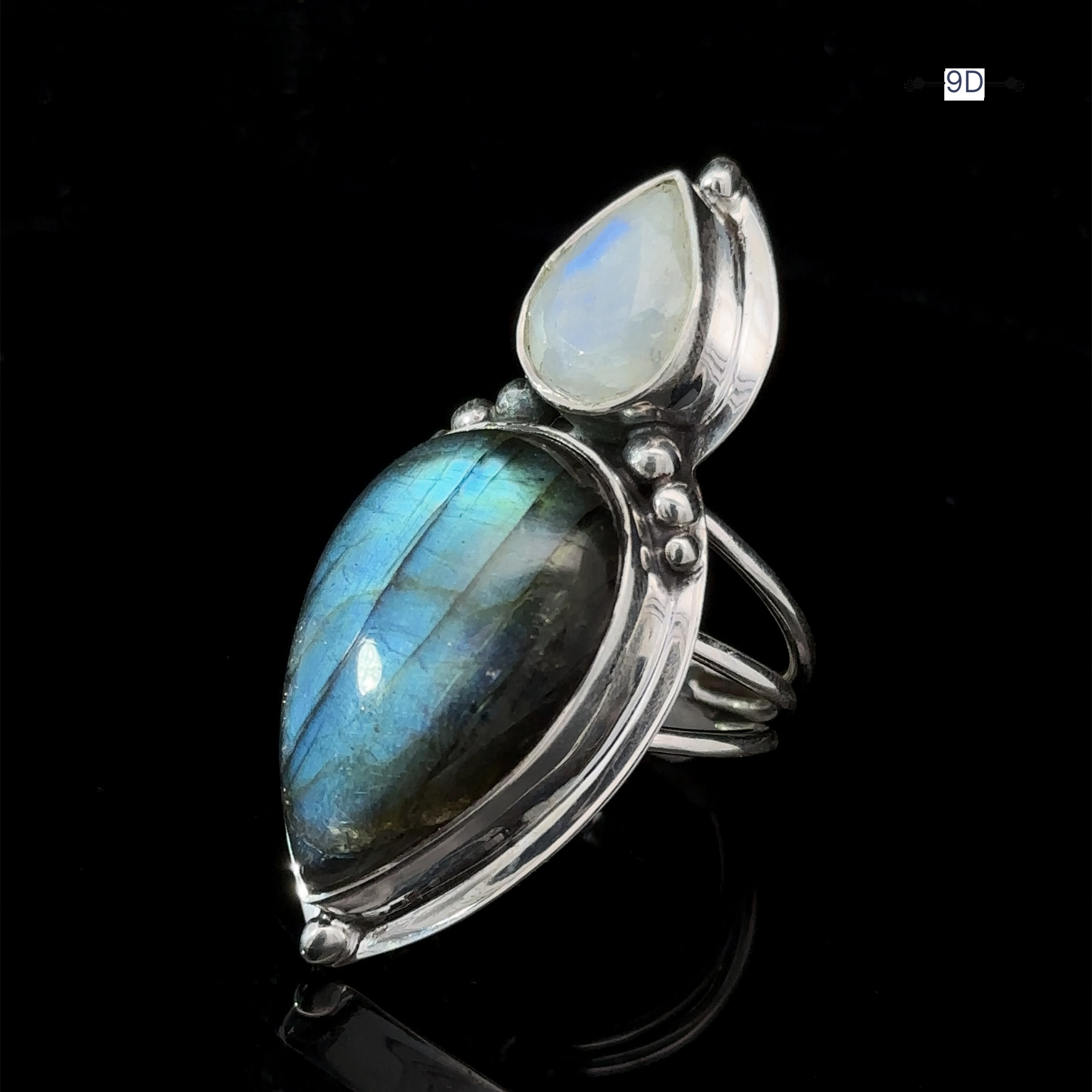 The Heavy Labradorite And Moonstone Ring showcases a large teardrop-shaped blue-green labradorite gemstone alongside a smaller white moonstone, all set against a striking black background.