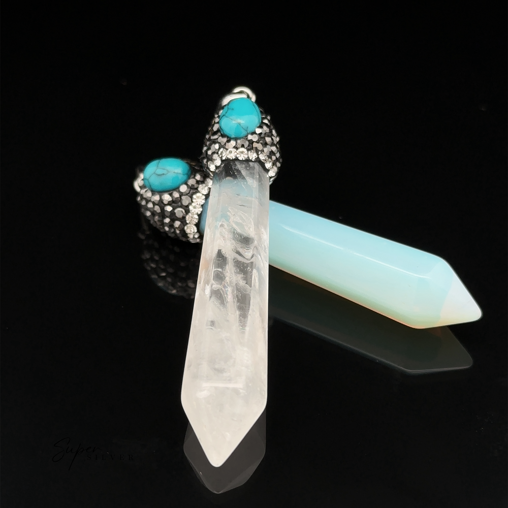 
                  
                    Two Stone Obelisk Pendants with pointed ends, one clear and one light blue, are attached to metal caps adorned with turquoise stones and hematite beads on a black background.
                  
                