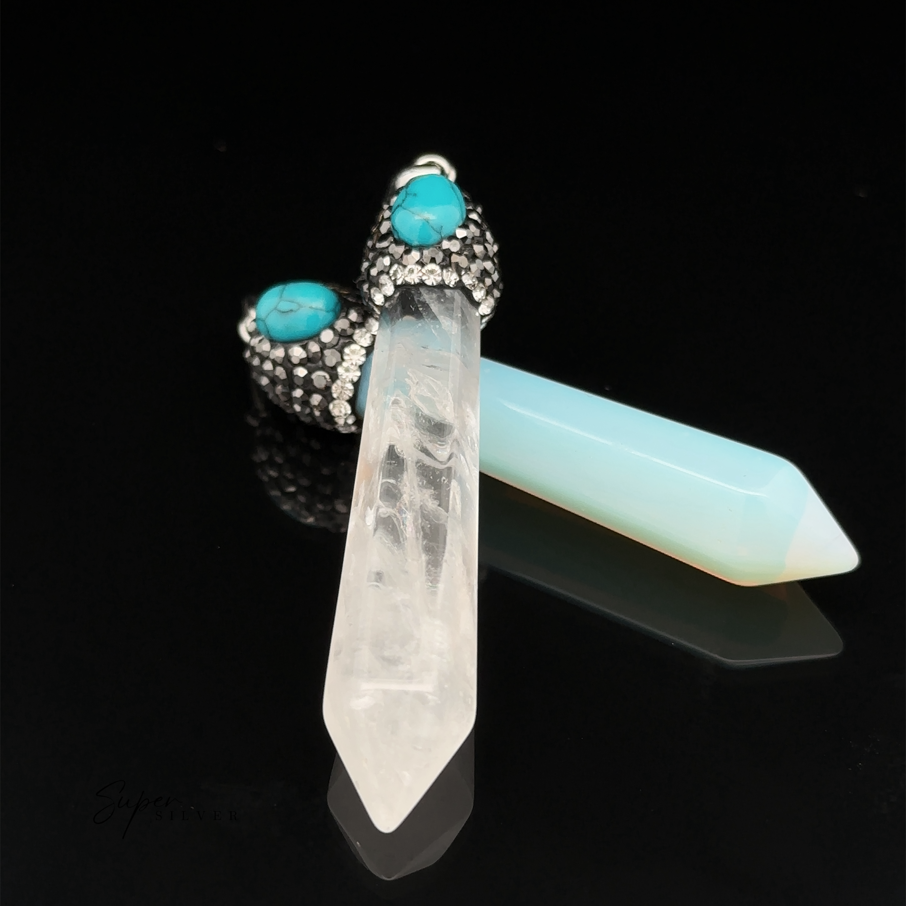 Two Stone Obelisk Pendants with pointed ends, one clear and one light blue, are attached to metal caps adorned with turquoise stones and hematite beads on a black background.
