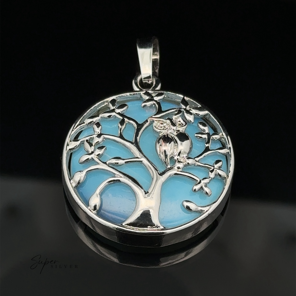 
                  
                    A round silver Owl and Tree Pendant depicting a tree with an owl perched on one of its branches against a blue background, crafted with mixed metals for added depth and character.
                  
                