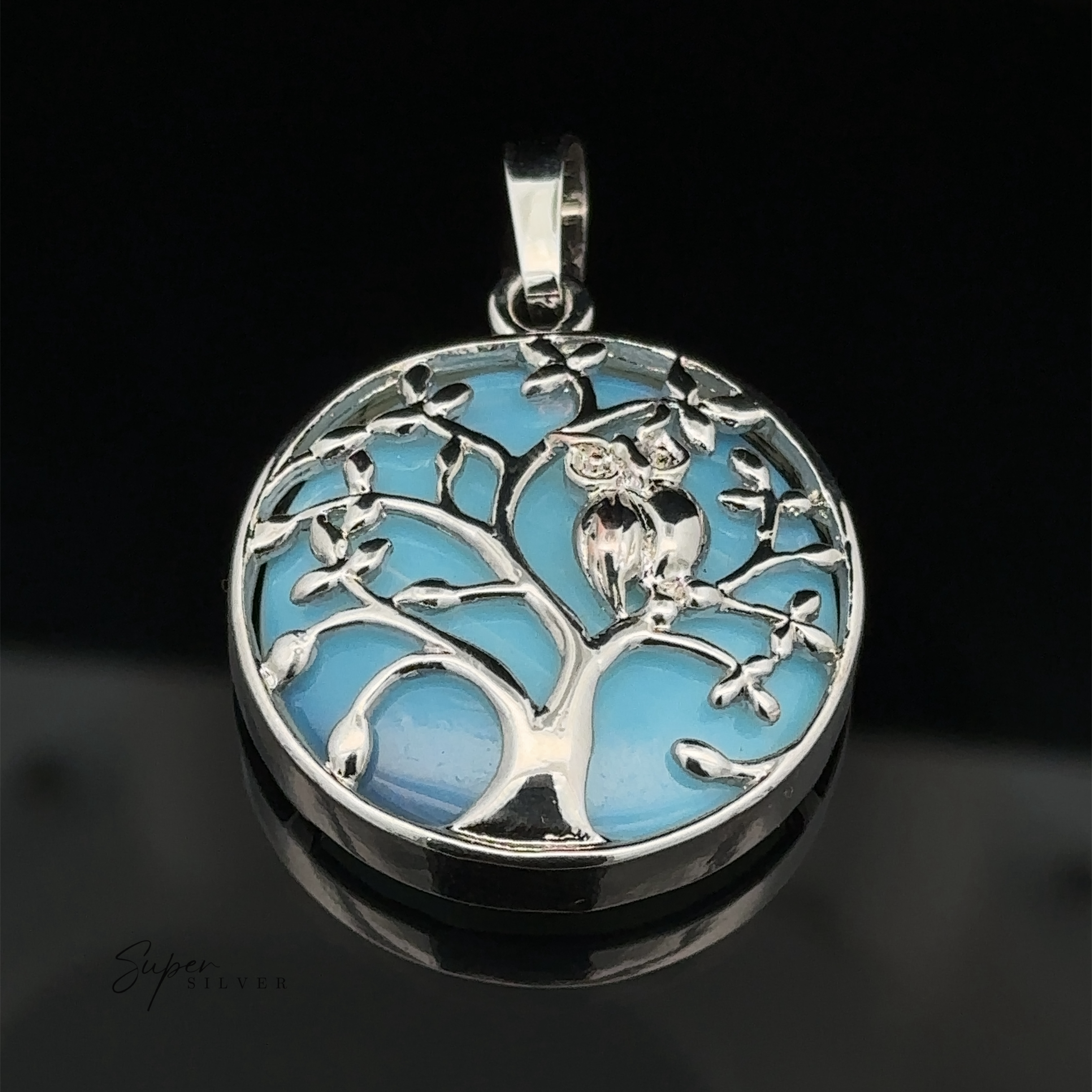 A round silver Owl and Tree Pendant depicting a tree with an owl perched on one of its branches against a blue background, crafted with mixed metals for added depth and character.