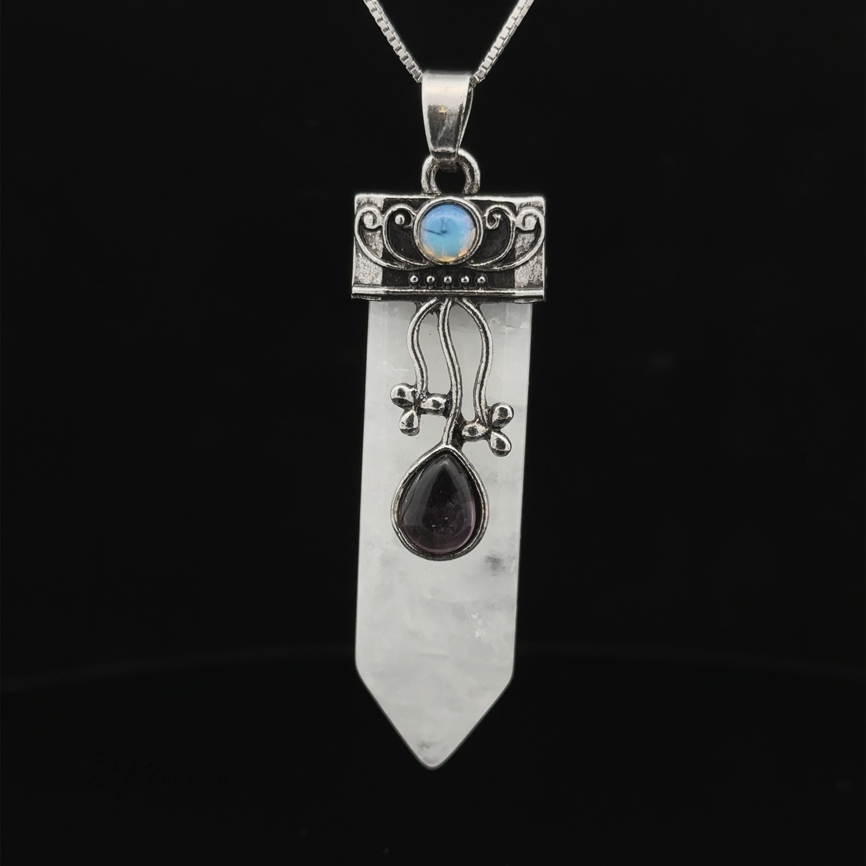 An Obelisk Crystal Stone Pendant with intricate silver designs features a blue gemstone, a pointed clear crystal, and a teardrop-shaped dark gem. Attached to a chain, the pendant stands out beautifully against a black background.