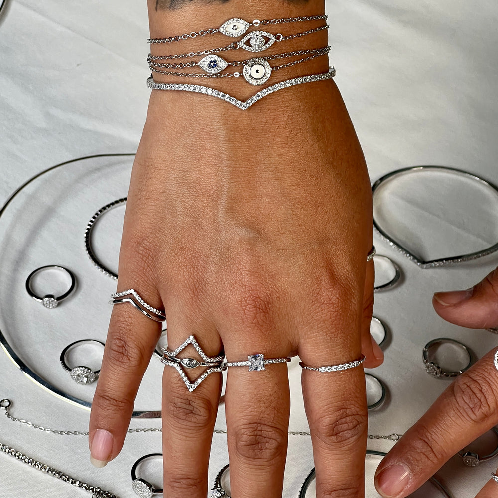 
                  
                    A hand with rings and layered bracelets, including a Cubic Zirconia Chevron Latch Bracelet and other rhodium plated sterling silver pieces, is displayed on a white surface among various elegant jewelry.
                  
                