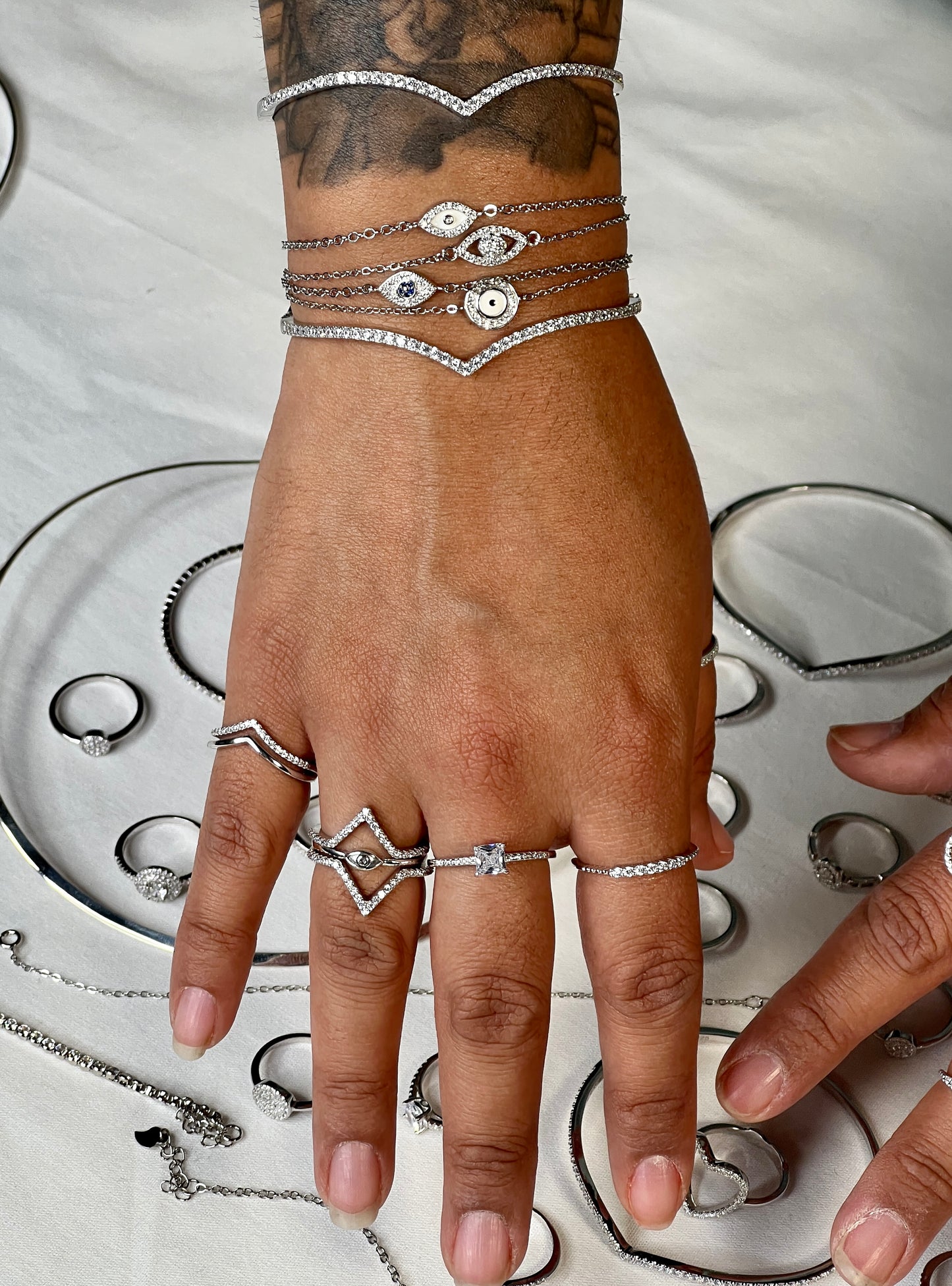 
                  
                    A hand with rings and layered bracelets, including a Cubic Zirconia Chevron Latch Bracelet and other rhodium plated sterling silver pieces, is displayed on a white surface among various elegant jewelry.
                  
                