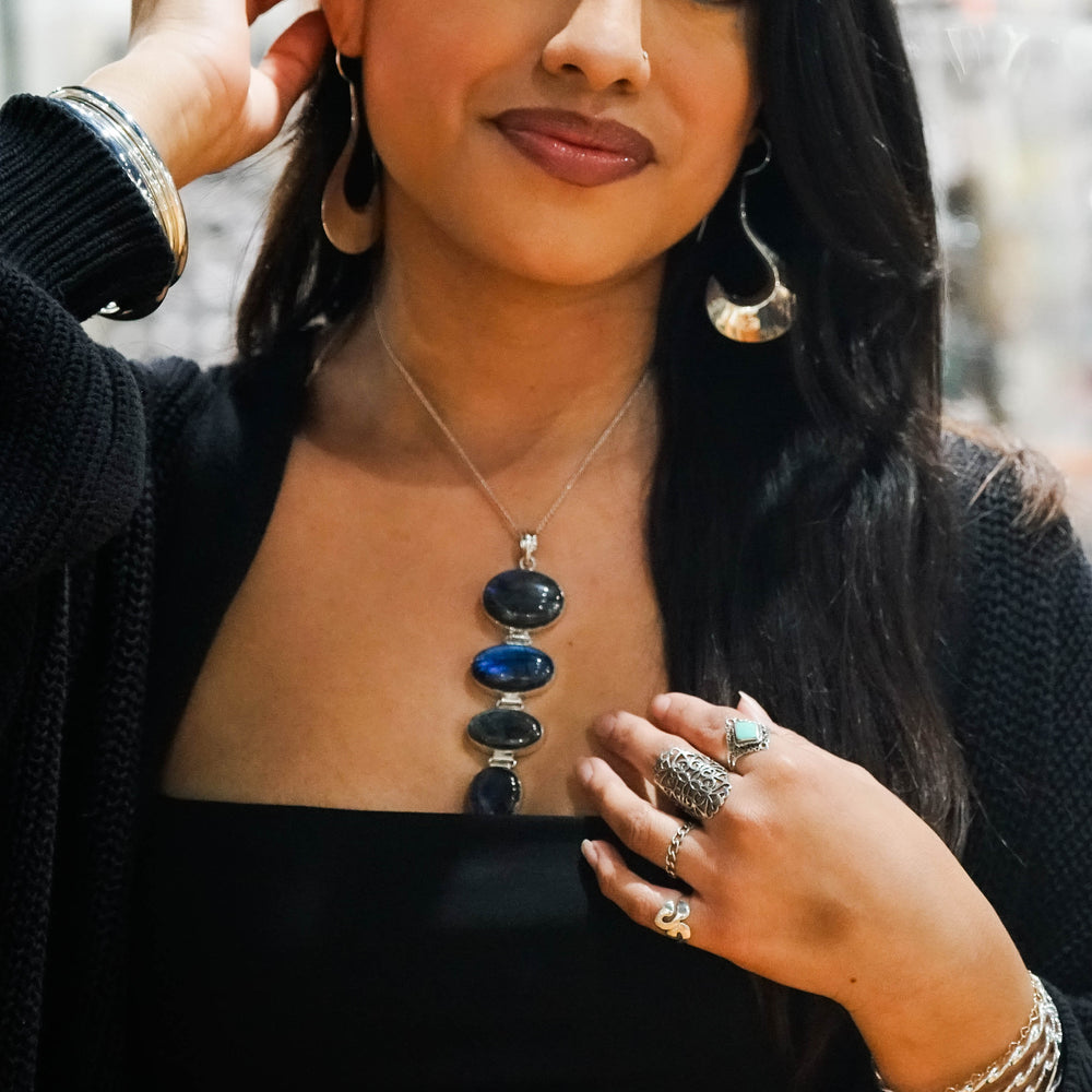 
                  
                    A woman wearing large hoop earrings, multiple rings, and a Tiered Statement Labradorite Pendant adjusts her earring while looking at the camera.
                  
                