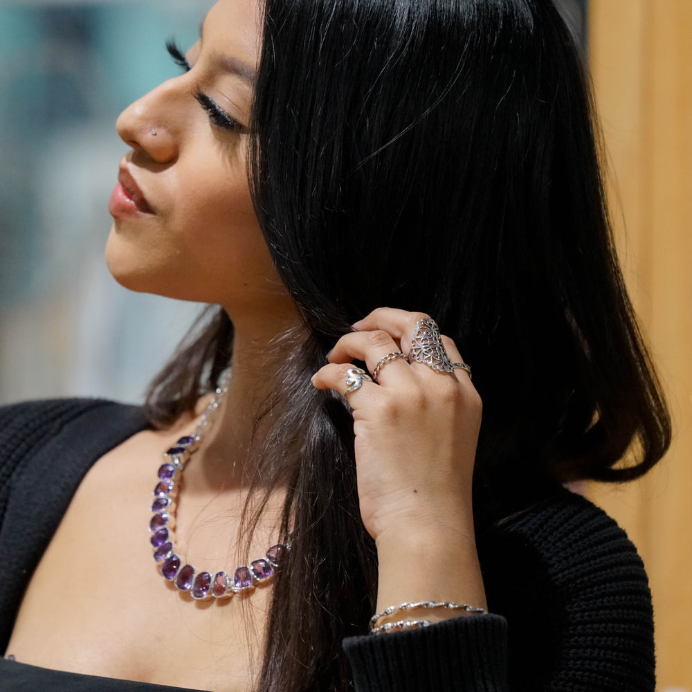 
                  
                    A woman with long dark hair, wearing a black top and a Statement Gemstone Necklace adorned with amethyst beads, rings, and a nose stud, holds a strand of her hair while looking to the side.
                  
                