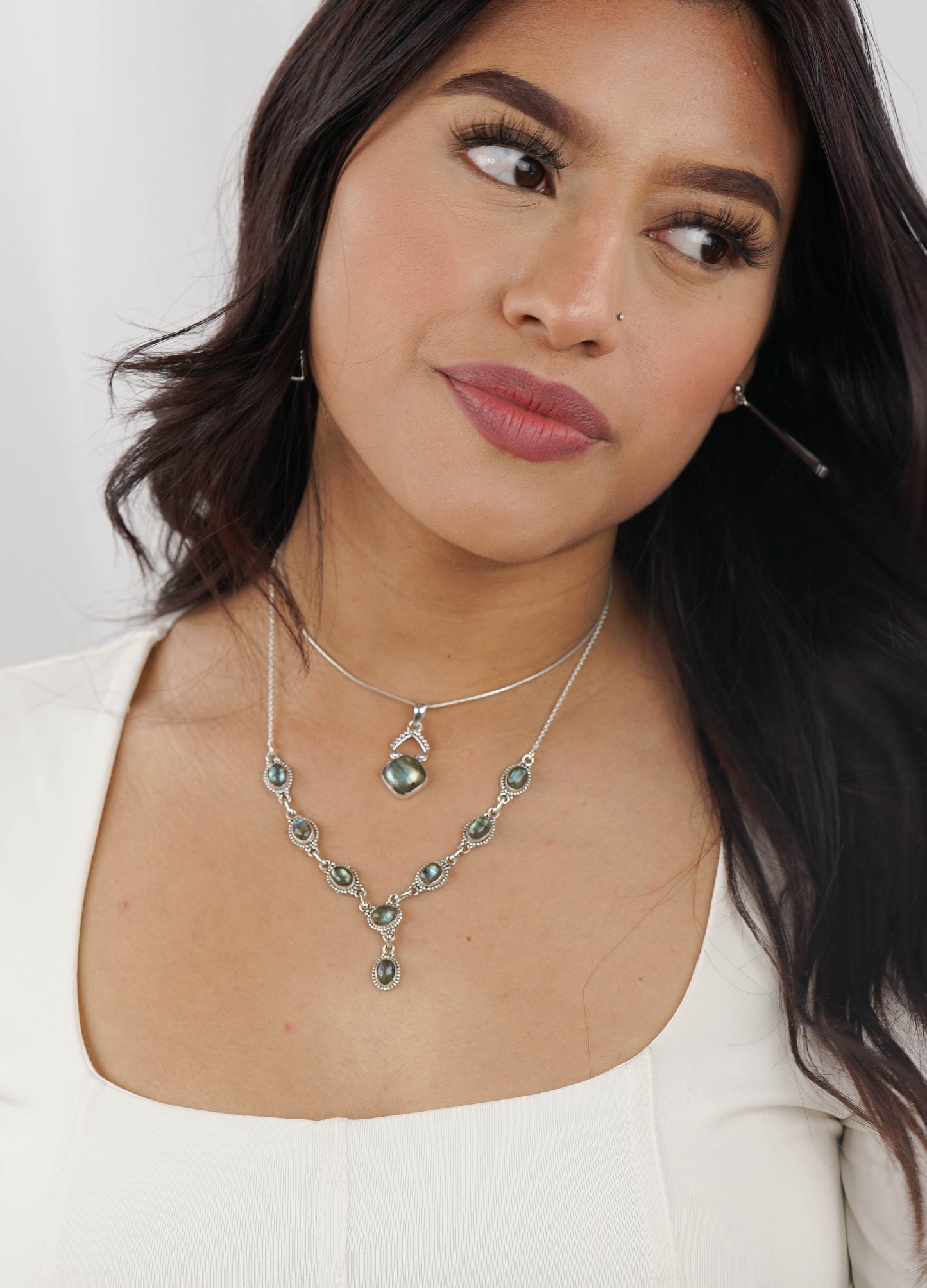 
                  
                    A woman with long, dark hair wears a white top and the Round Gemstone Y Necklace with Ball Border featuring green gemstones. She gazes to the side against a neutral background, exuding boho-chic charm.
                  
                