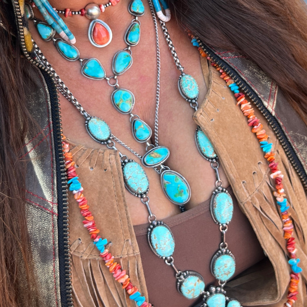 
                  
                    Close-up of a person wearing the Stunning Sonora Gold Lariat Necklace And Earrings Set adorned with authentic Native American turquoise stones, coral beads, and silver details, paired with a brown fringed suede jacket.
                  
                