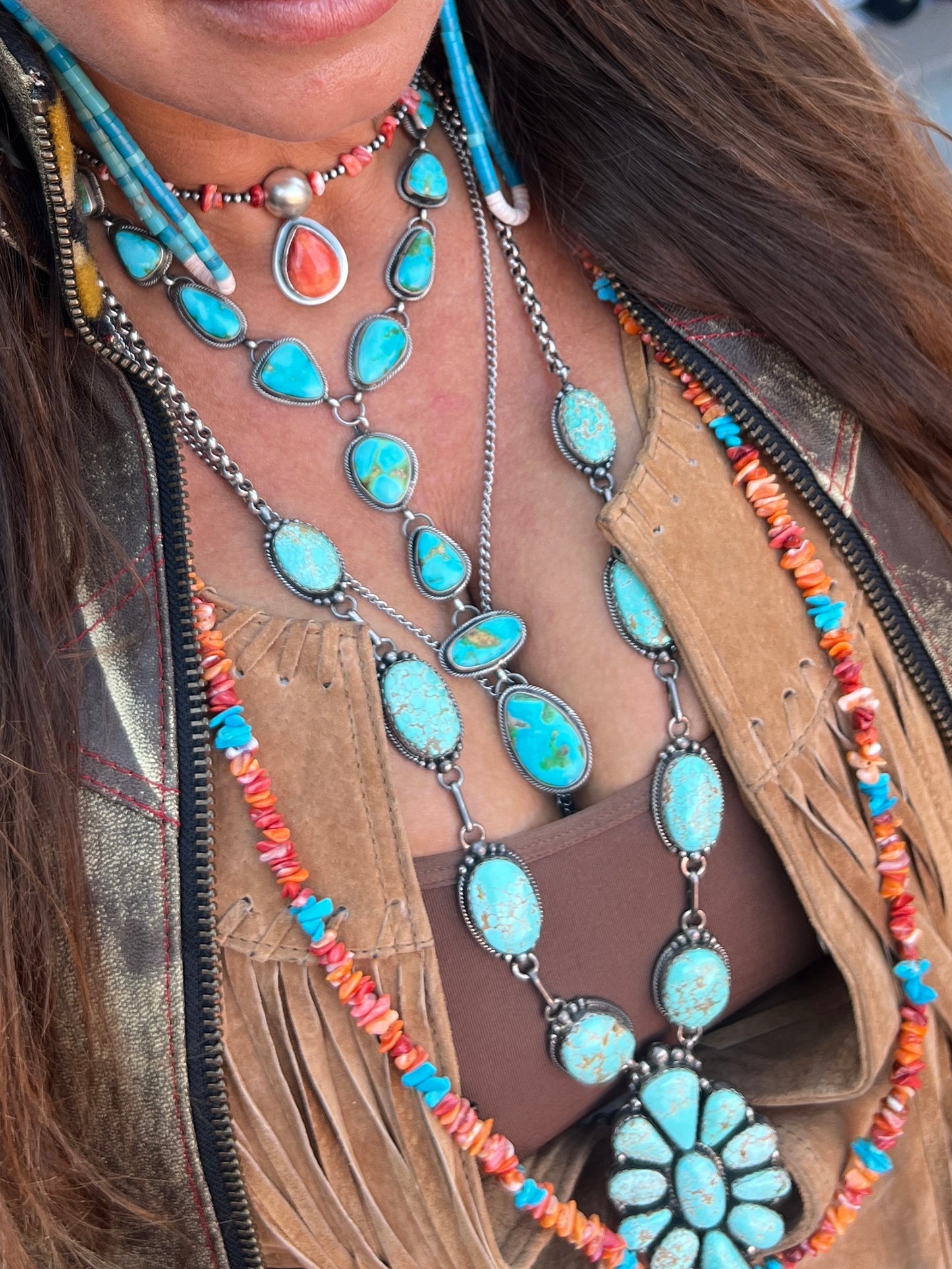 
                  
                    Close-up of a person wearing the Stunning Sonora Gold Lariat Necklace And Earrings Set adorned with authentic Native American turquoise stones, coral beads, and silver details, paired with a brown fringed suede jacket.
                  
                