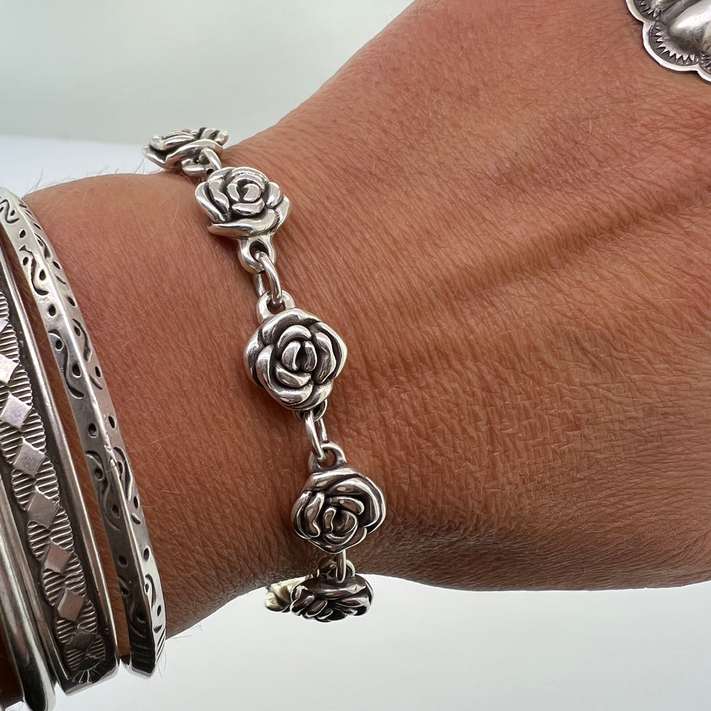 
                  
                    A wrist adorned with three silver bracelets, including a Chic Link Rose Bracelet with vintage vibes and another with intricate patterns. The thumb is partially visible wearing a large, detailed .925 sterling silver ring.
                  
                