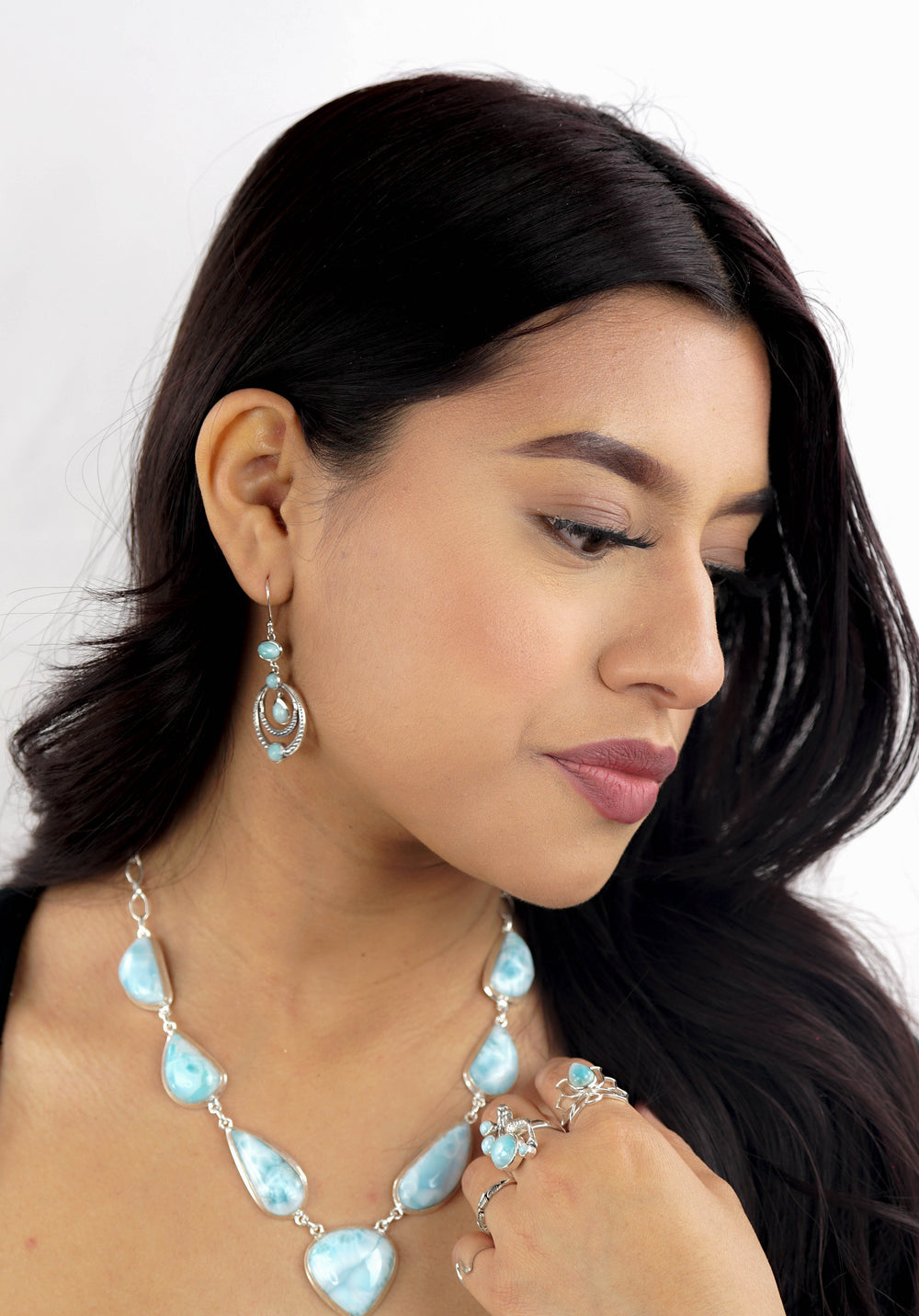 A woman with long, dark hair is shown wearing turquoise jewelry, including earrings, rings, and a Beautiful Larimar Necklace With Teardrop Shape Stones. She is facing slightly to the side against a white background.