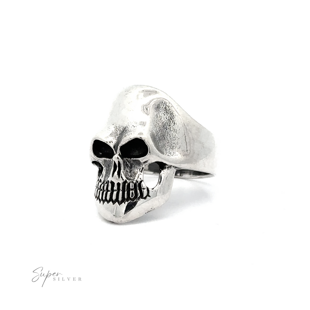 A silver ring shaped like a detailed human skull with hollow eyes and teeth is photographed against a white background. "Silver Skull Statement Ring" is written in small text at the bottom left corner, showcasing the simple skull statement ring's edgy elegance in .925 Sterling Silver.