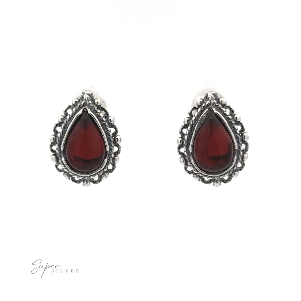 
                  
                    A pair of Framed Teardrop Amber Studs featuring dark red gemstones bordered with intricate silver detailing, displayed on a white background, exuding a sense of vintage romance.
                  
                