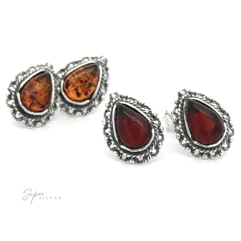 Two pairs of Framed Teardrop Amber Studs with ornate silver borders, one set featuring Baltic Amber and the other deep red stones, exude vintage romance as they're elegantly displayed on a white background.