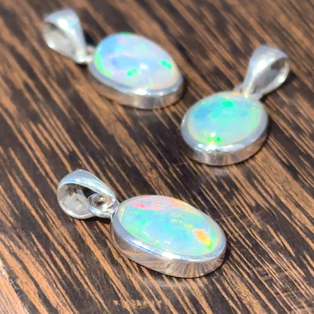 
                  
                    Three Oval Ethiopian Opal Pendants in sterling silver settings are elegantly displayed on a wooden surface, embodying a minimalist style.
                  
                