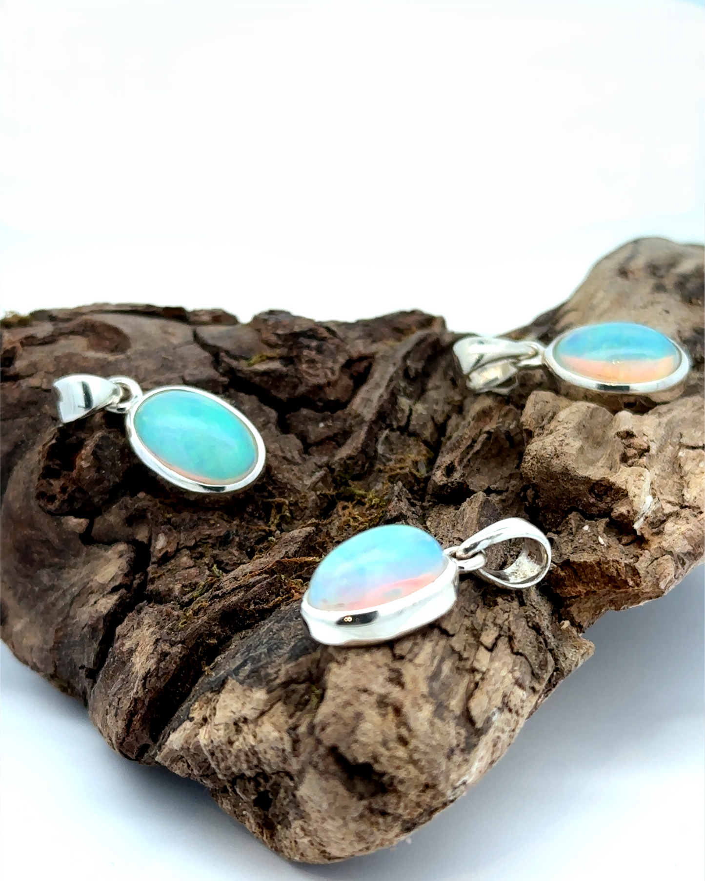 
                  
                    Three Oval Ethiopian Opal Pendants with sterling silver settings are elegantly displayed on a piece of driftwood, embodying a minimalist style.
                  
                