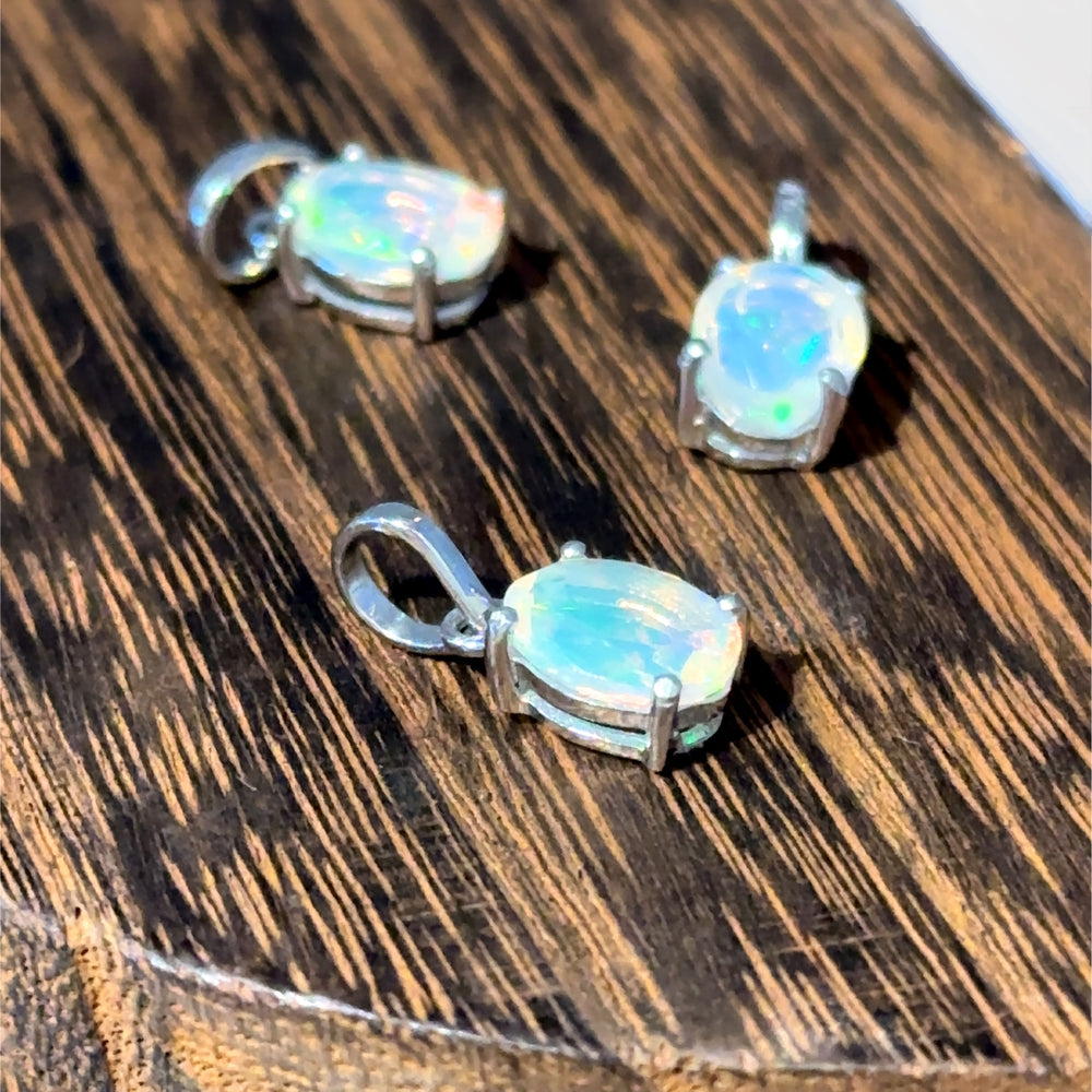 
                  
                    Three Tiny Oval Pronged Ethiopian Opal Pendants are set in sterling silver, resting gracefully on a wooden surface.
                  
                