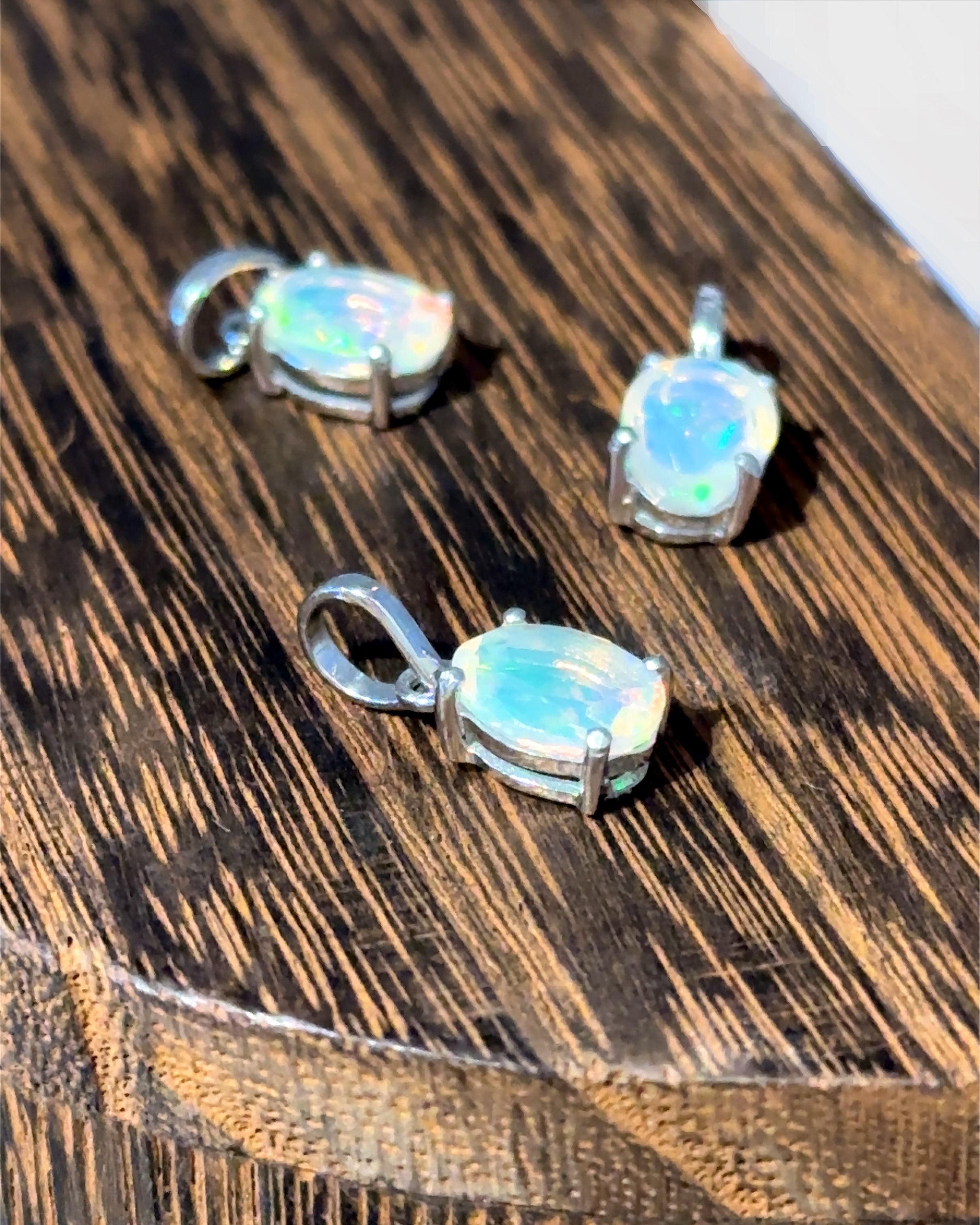 Three Tiny Oval Pronged Ethiopian Opal Pendants are set in sterling silver, resting gracefully on a wooden surface.