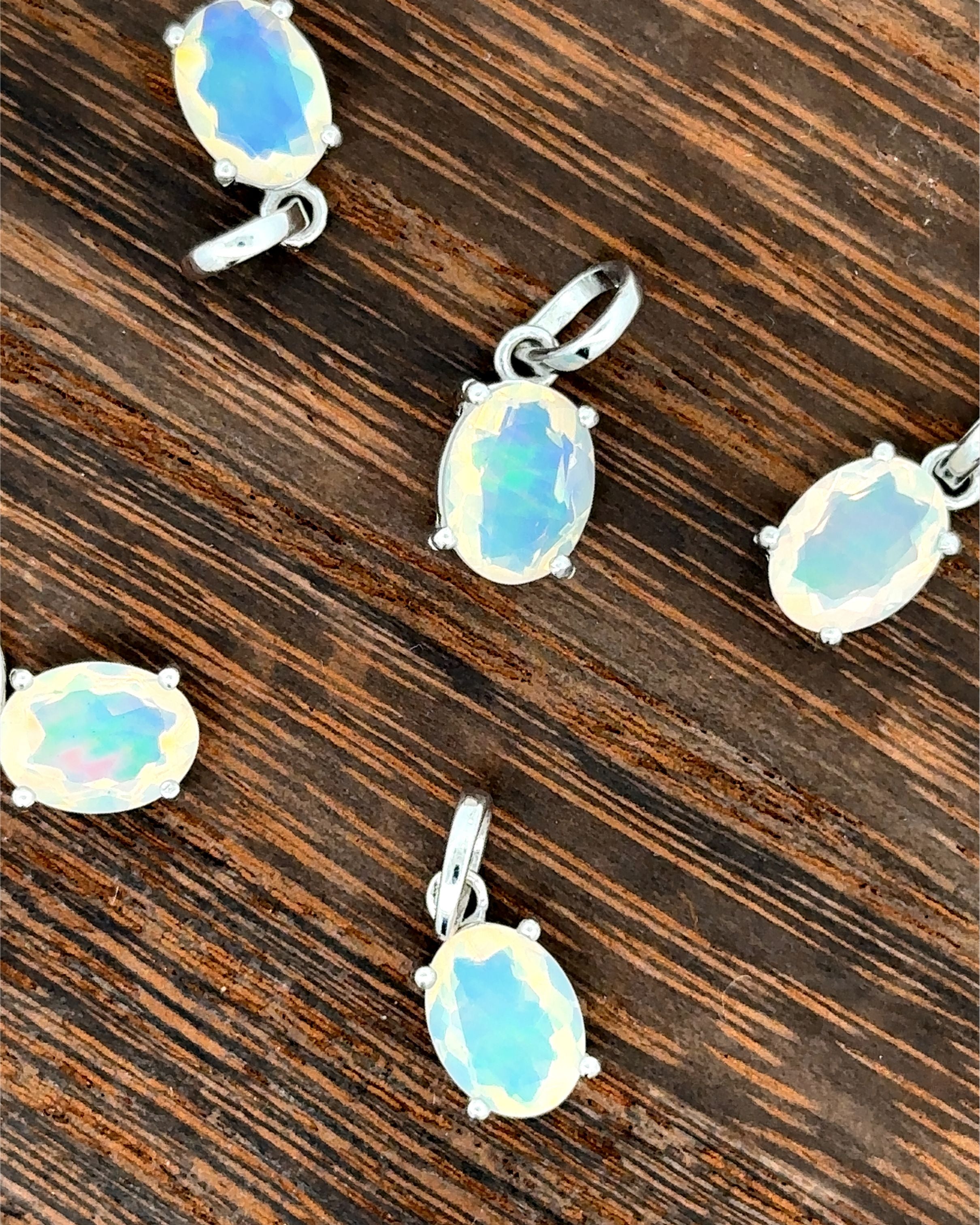 Five Tiny Oval Pronged Ethiopian Opal Pendants, crafted with Ethiopian opal and set in sterling silver, are elegantly arranged on a wooden surface.