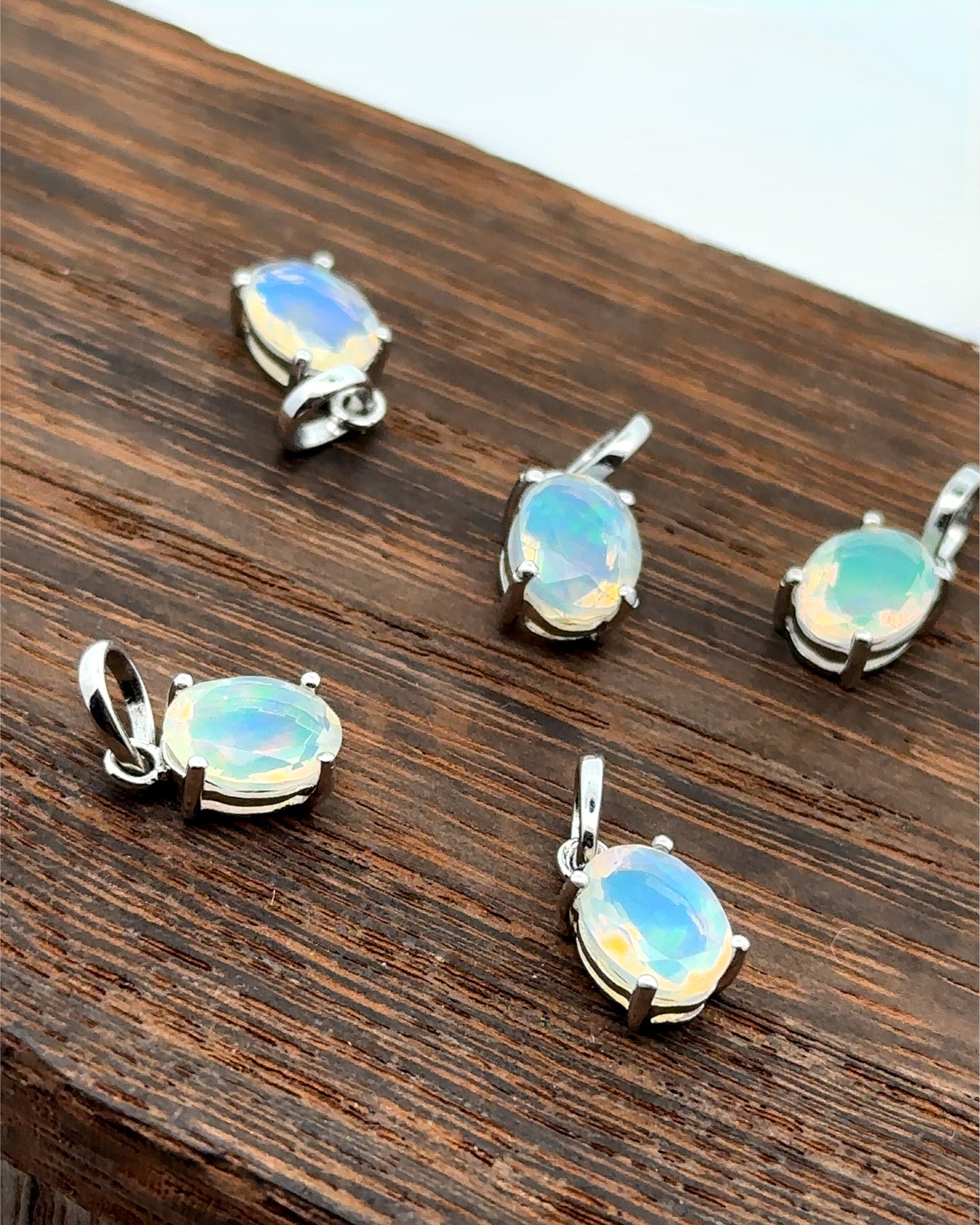 
                  
                    Five Tiny Oval Pronged Ethiopian Opal Pendants, each crafted with Ethiopian opal set in sterling silver, are elegantly displayed on a wooden surface.
                  
                