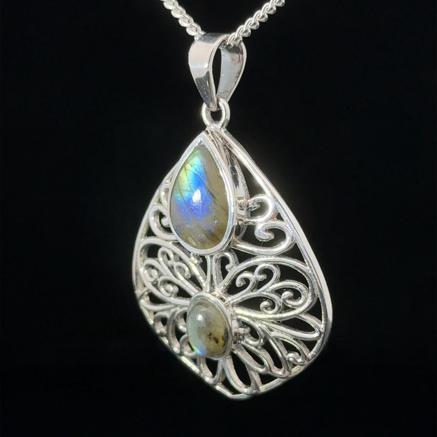 
                  
                    A Teardrop Filigree Gemstone Pendants with an intricate design featuring two labradorite stones, one teardrop-shaped and one oval-shaped, hangs from a silver chain against a black background. An amethyst accent adds a touch of mystique.
                  
                