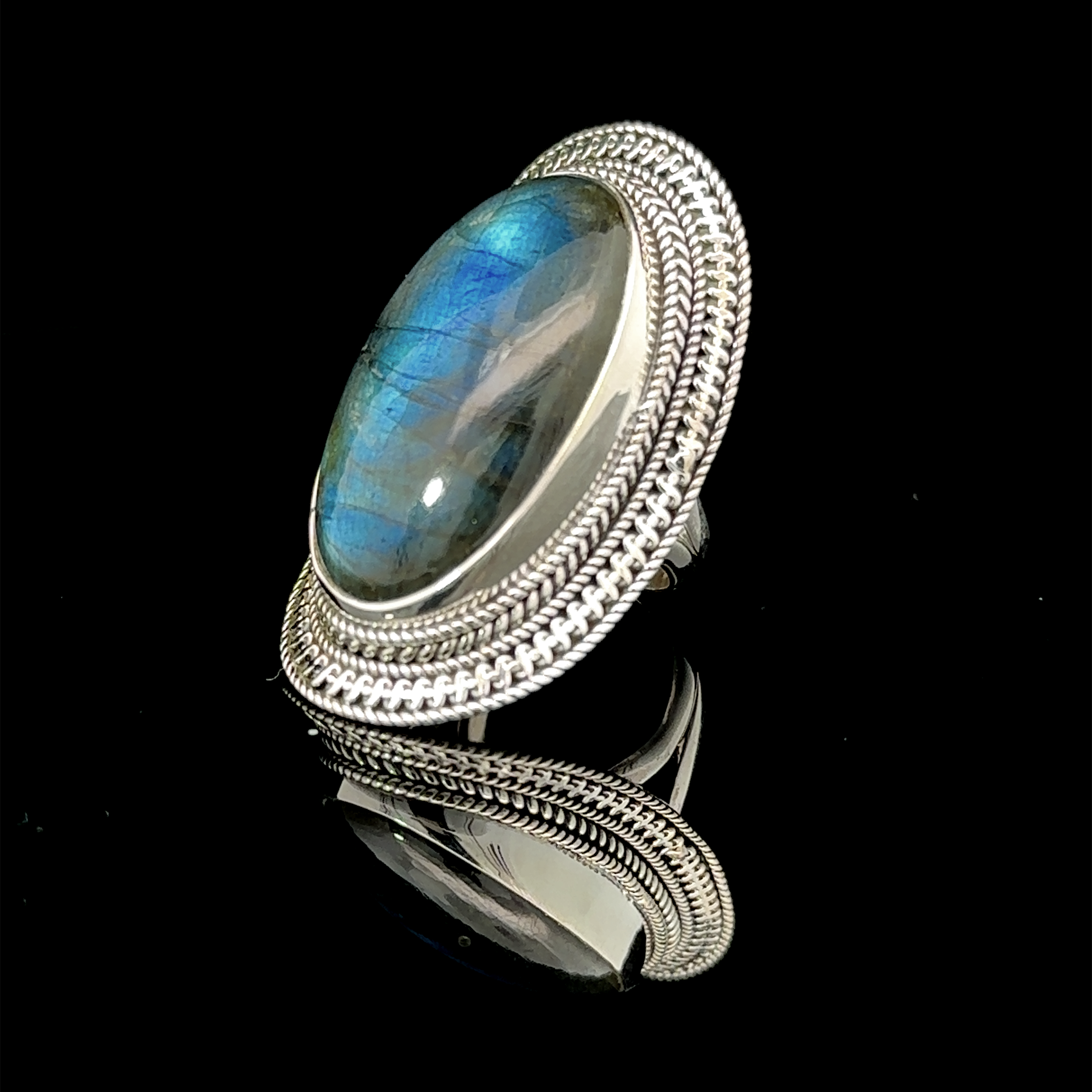 A Large Oval Shield Gemstone Ring with an oval labradorite stone featuring a blue and green sheen, set against a black background, exudes a bohemian flair.