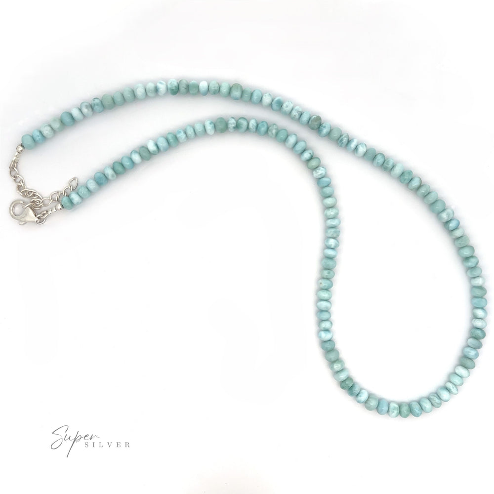 A "Larimar Beaded Necklace" made of round, light blue beads arranged in a single strand with a silver clasp. The background is white. "Super Silver" is written in small text at the bottom left corner.